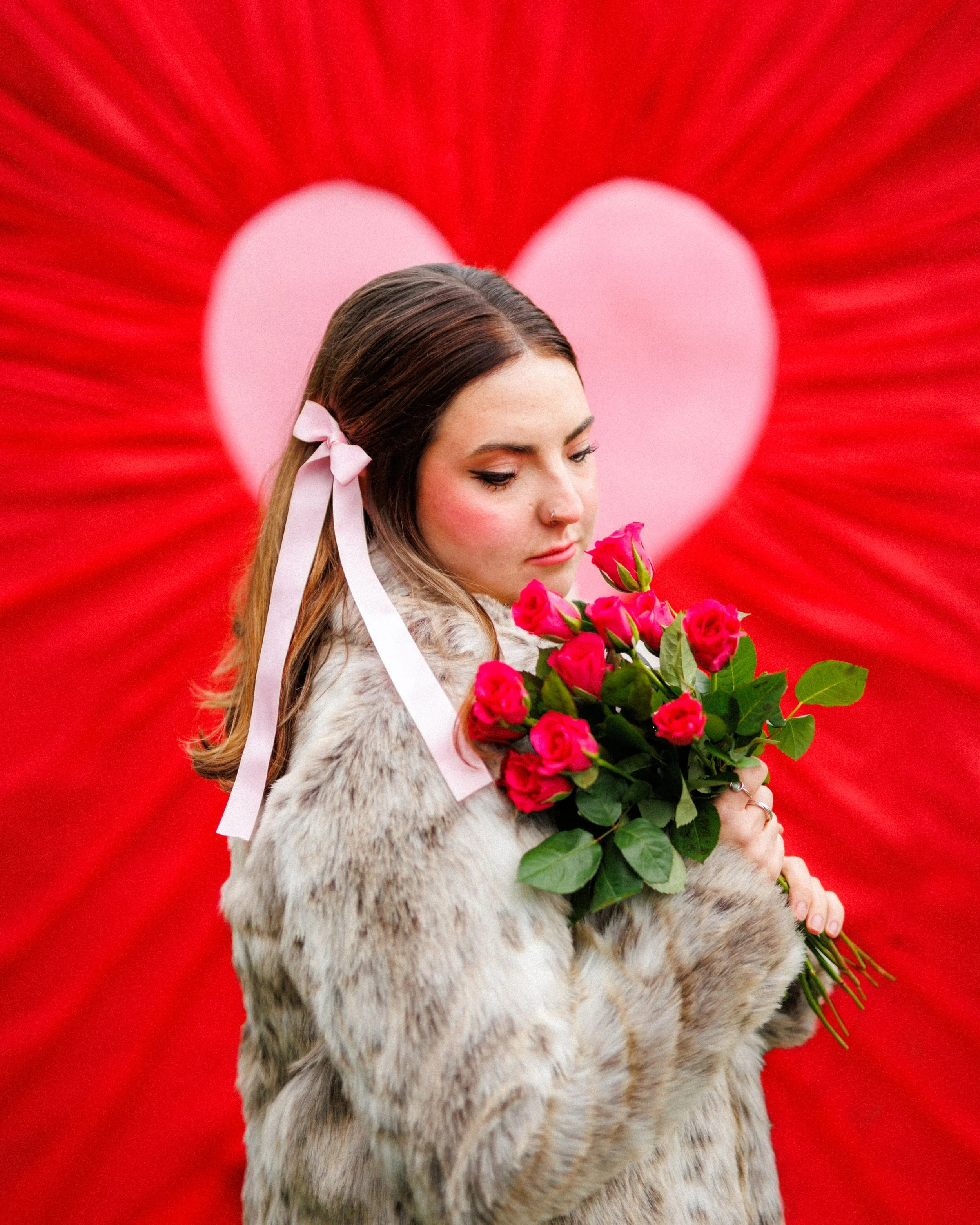 A woman with brown hair and a nose ring holding a bouquet of pink roses on Valentine's Day with a red background and a large pink heart