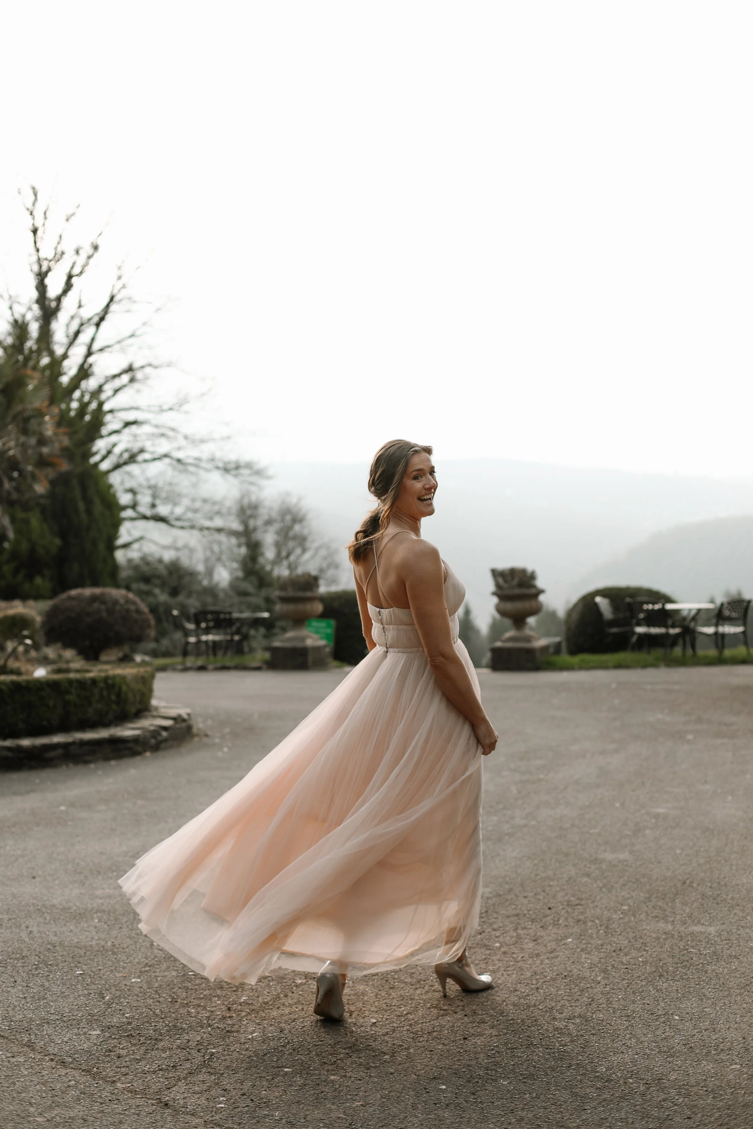 A woman in a flowing, light peach-colored dress and high heels is smiling and twirling outdoors on a gray paved surface, with a garden and distant mountains in the background.