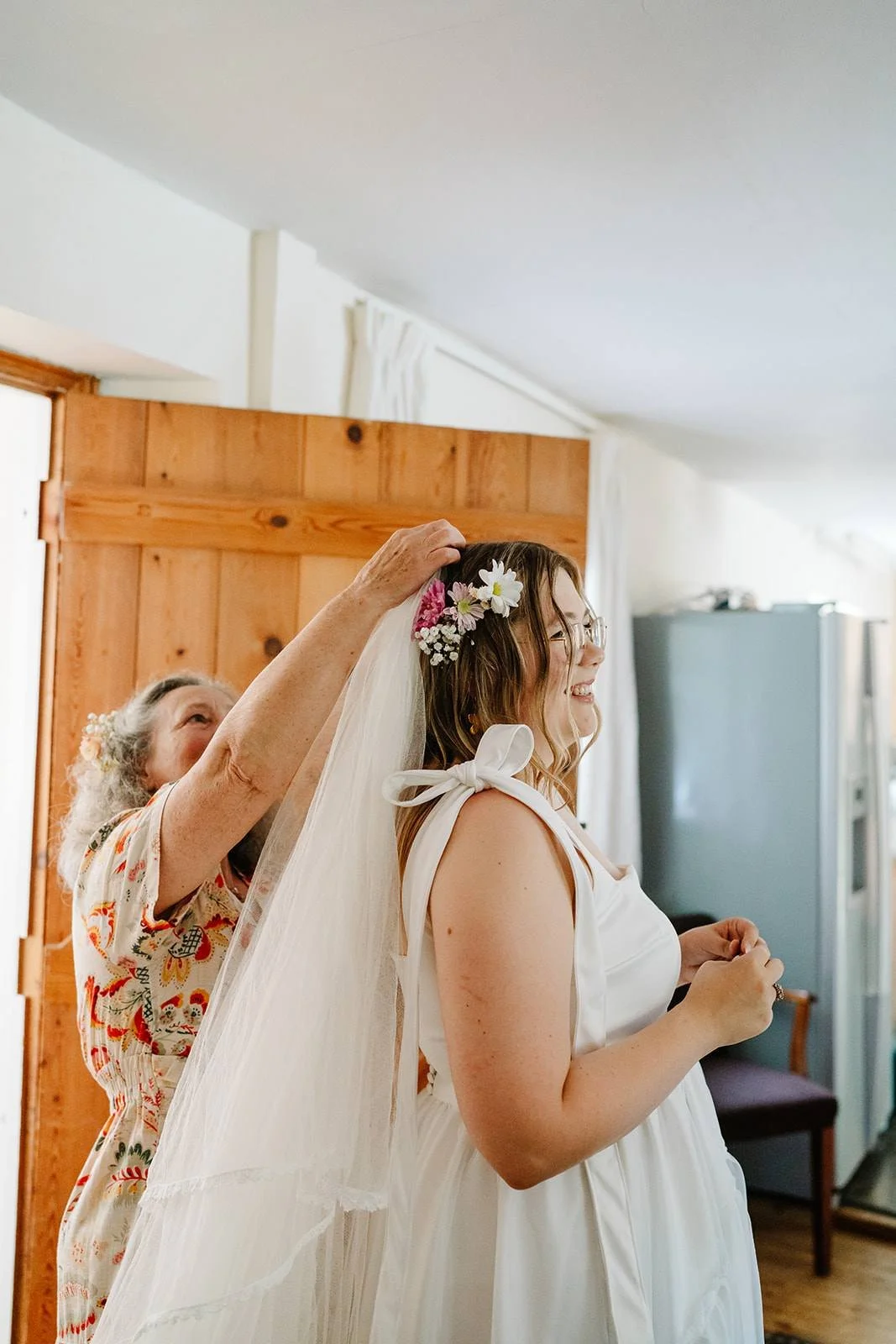 A young woman wearing glasses and a white dress is smiling as an older woman, likely her mother, helps her put on a wedding veil decorated with flowers in a cozy room.