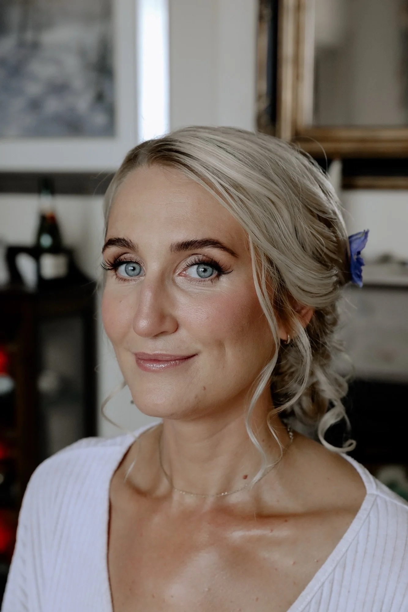 Close-up of a smiling woman with blonde hair and blue eyes, wearing a white top, with a blurred background.