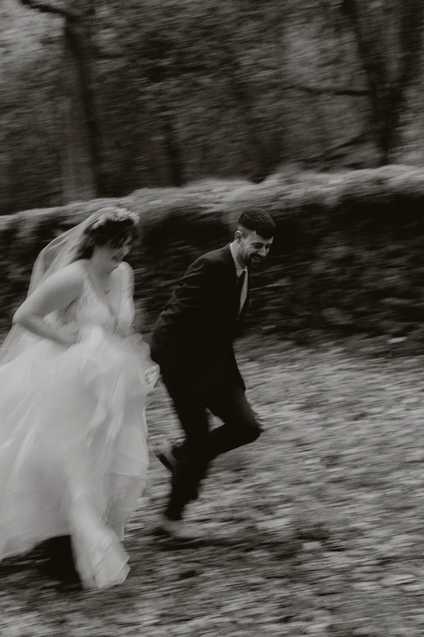 A couple dressed in wedding attire running outdoors, smiling, with a blurred background of trees and grass.