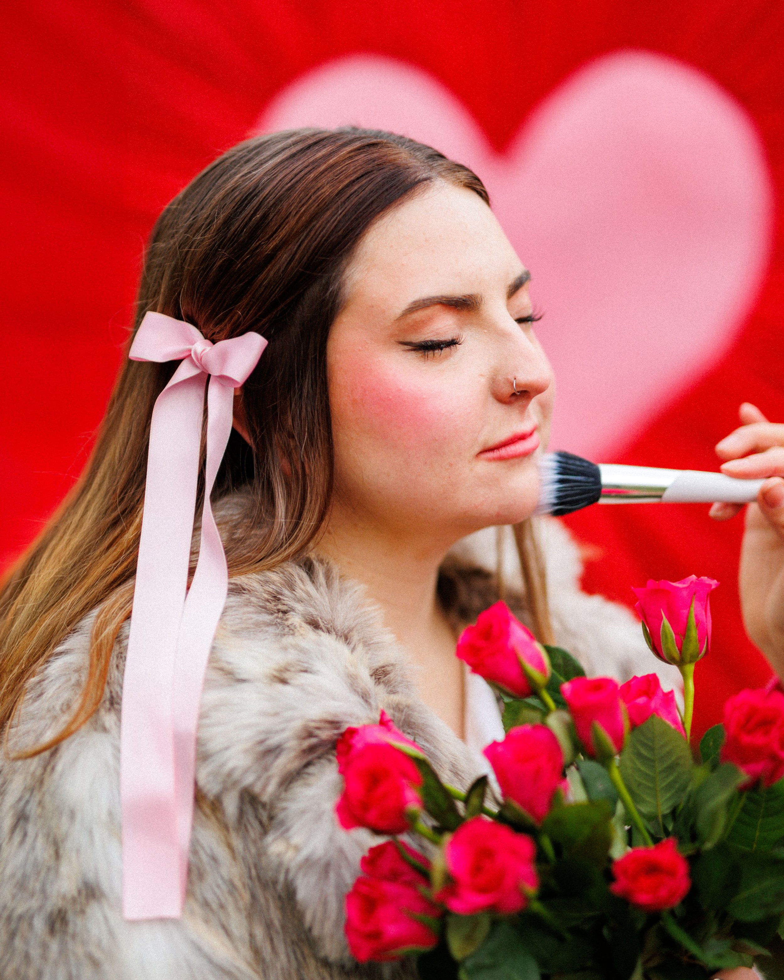 A woman with long brown hair and a pink ribbon tied in her hair is posing with a bouquet of pink roses while her face is being gently made up with a brush, with a large pink heart in the background.