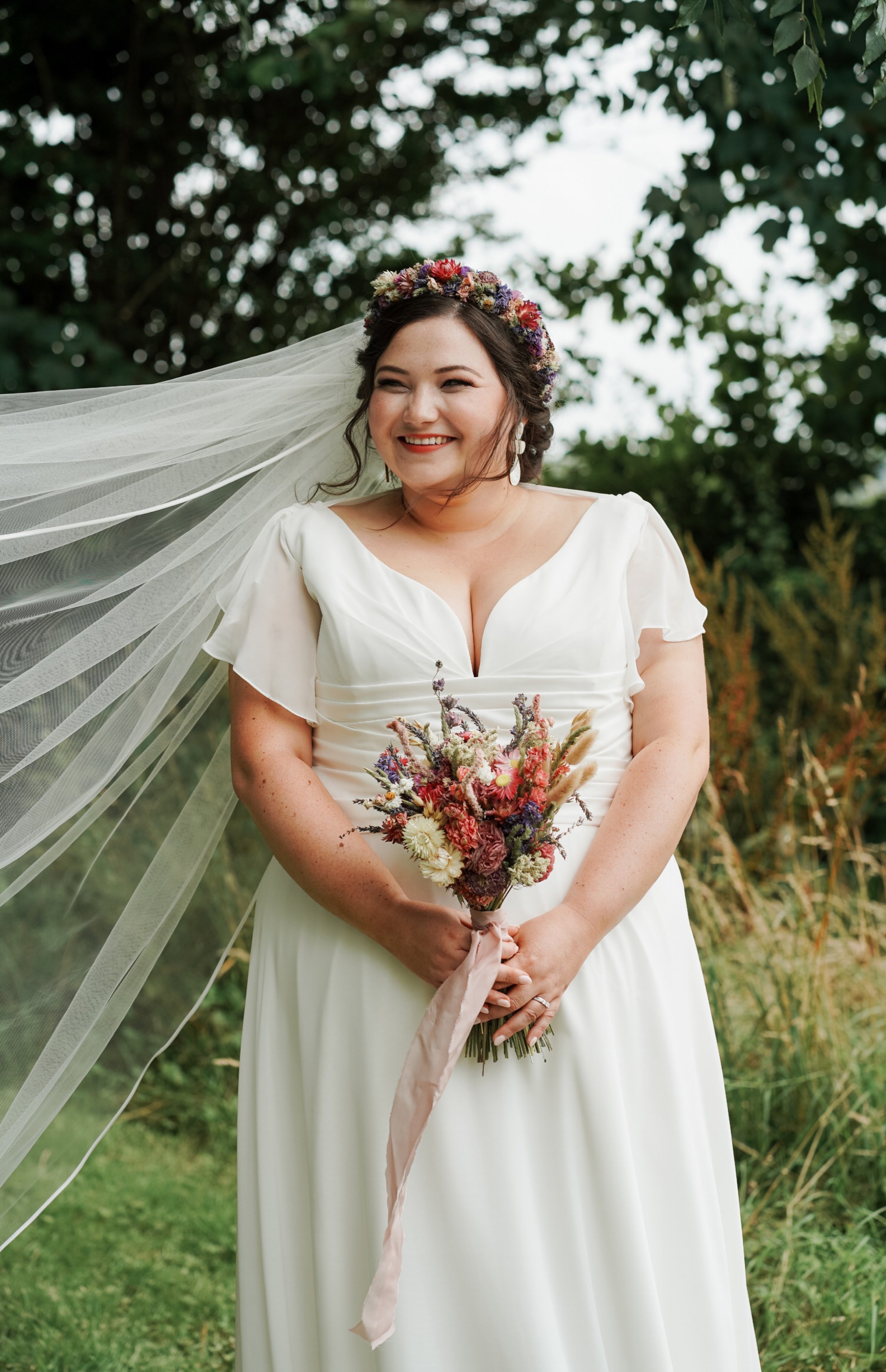 A smiling bride in a white wedding dress holding a bouquet of flowers outdoors at a wedding venue with greenery in the background, wearing a floral crown and a veil flowing in the wind.