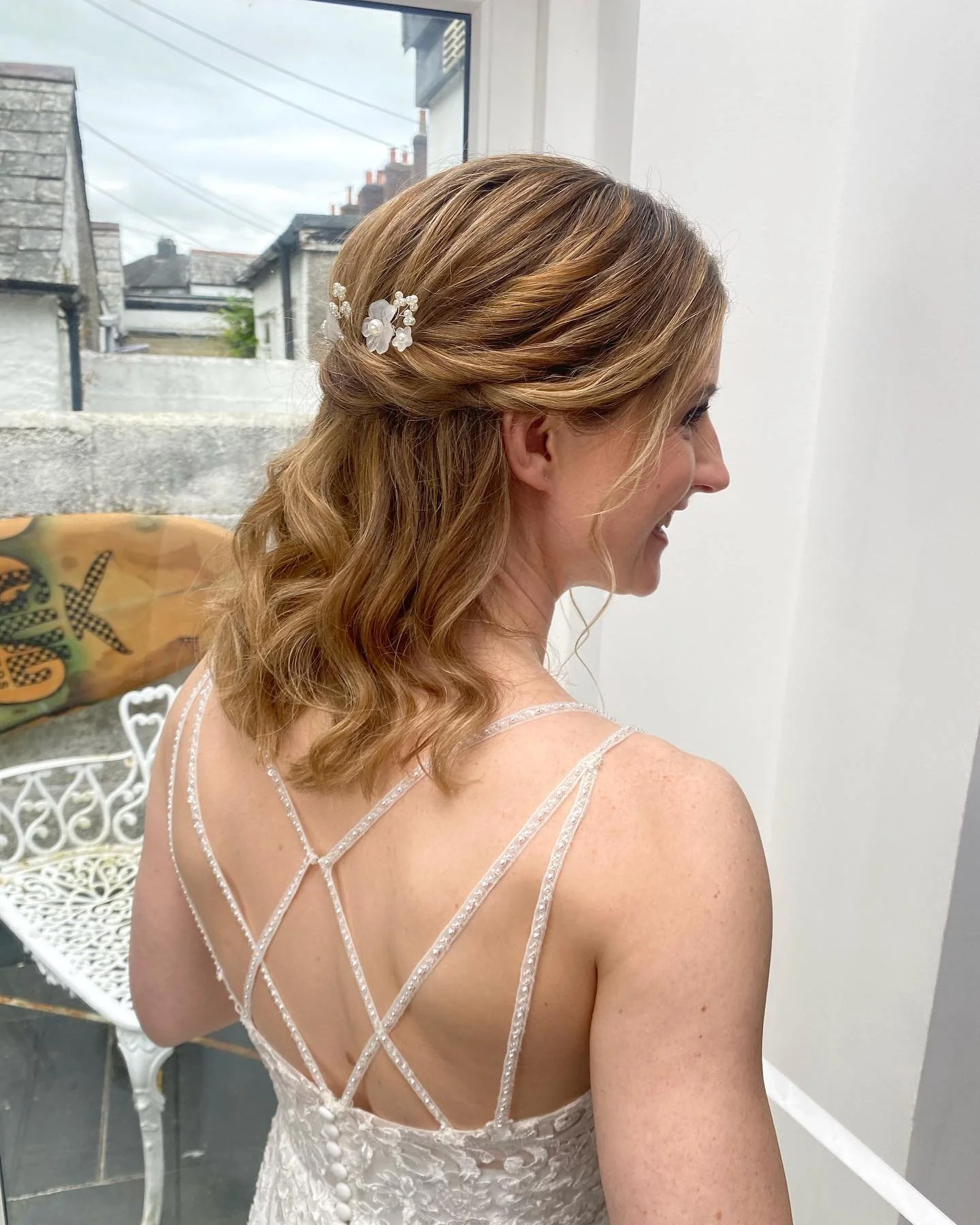 A woman with wavy, shoulder-length hair styled with a white floral hair clip, wearing a white lace dress with thin, crisscrossing straps on her back, smiling and looking to the right in a bright room with a window in the background.