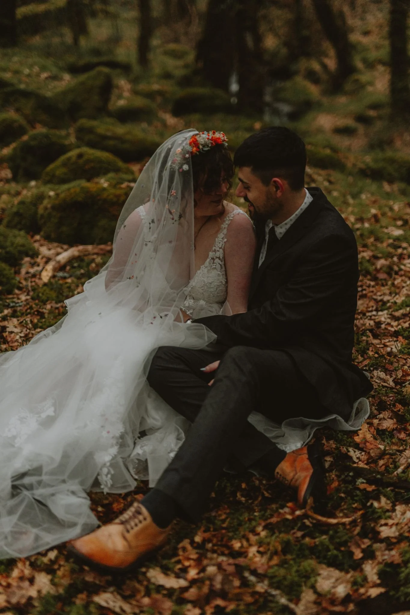 A bride in a white lace wedding dress with a veil and floral crown, and a groom in a black suit, sitting closely together on a forest floor covered with leaves and moss, smiling and leaning their foreheads together.