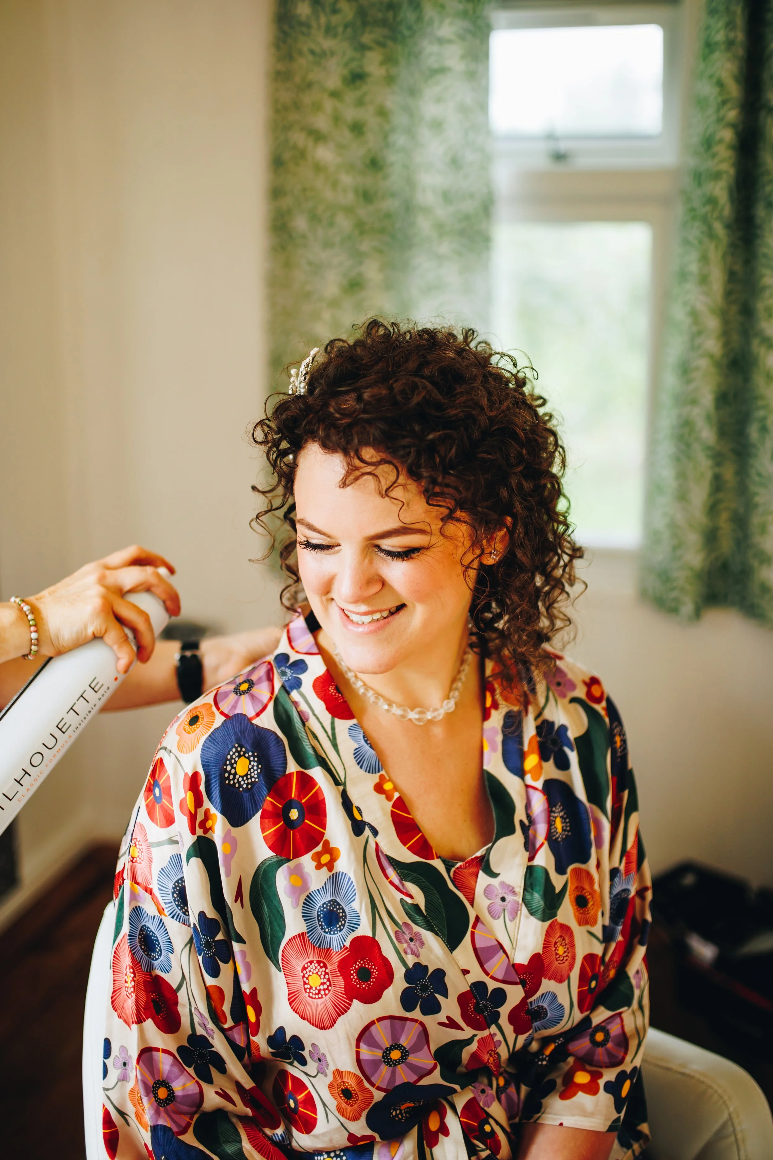A smiling woman with curly hair getting her hair styled with hair spray in a room with natural light and green curtains.