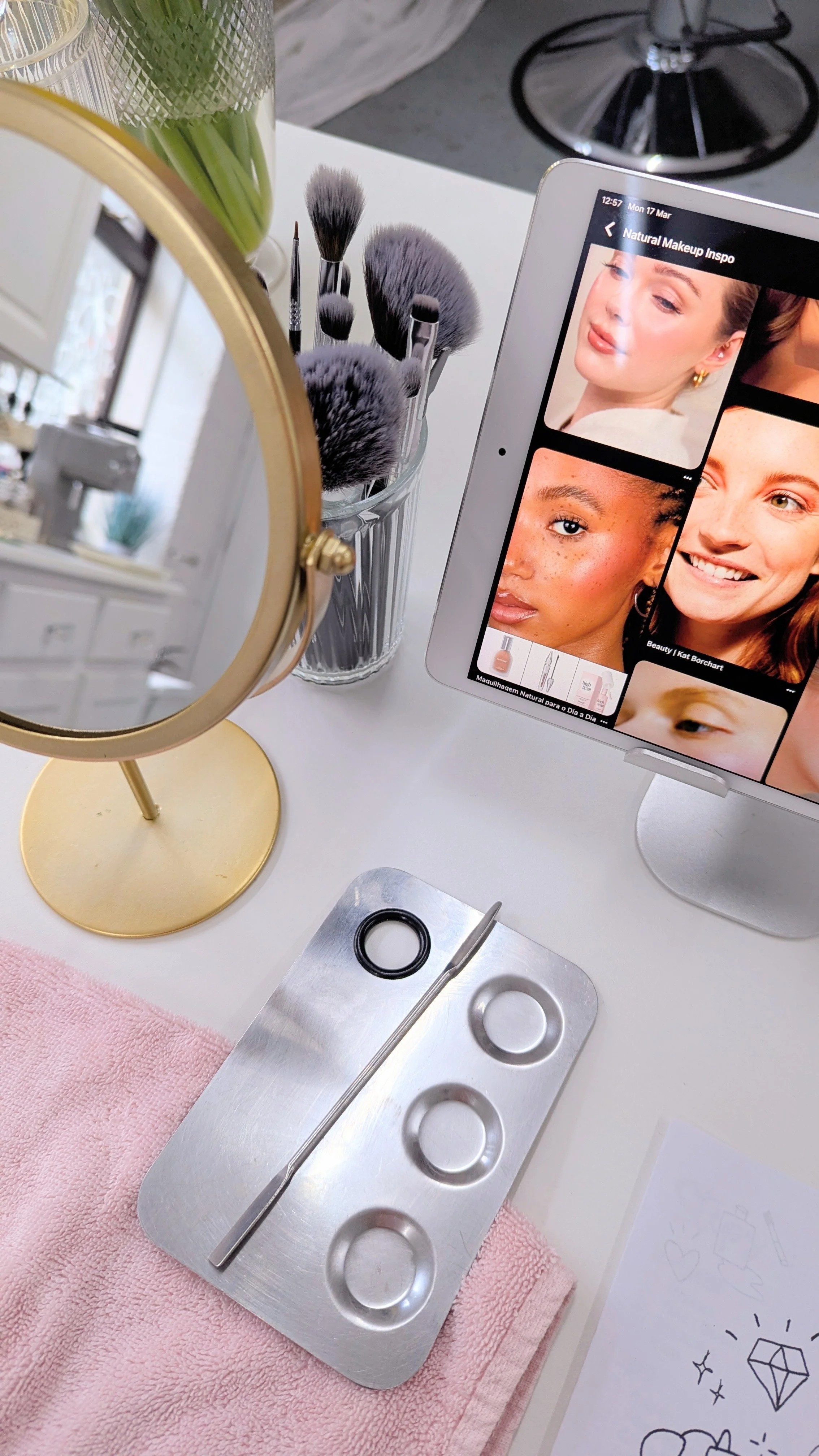 A vanity setup with makeup brushes in a glass container, a small mirror, a pink towel, and a tablet displaying makeup tutorial images. There is also a face chart drawing on paper.