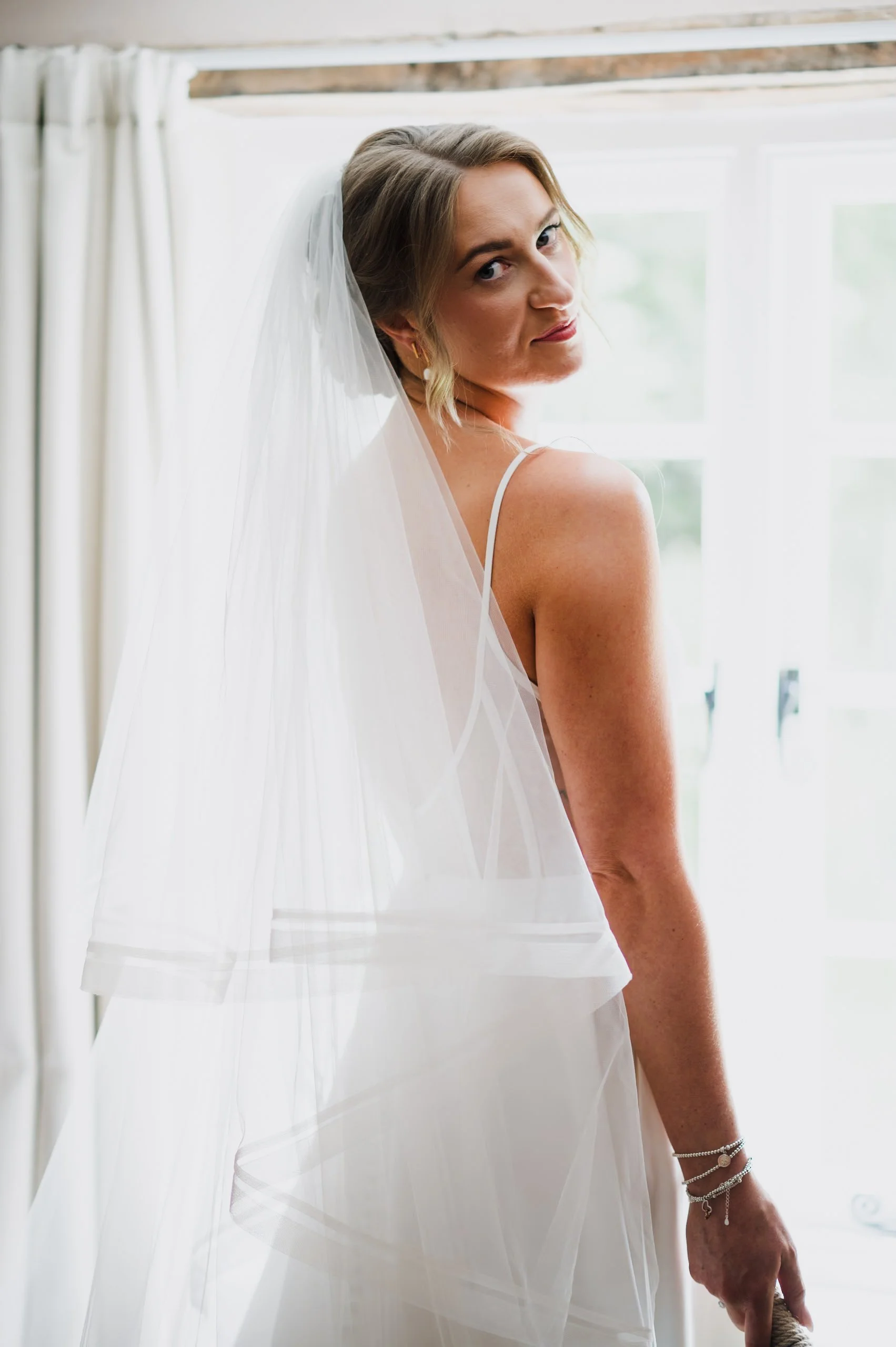 A bride in a white wedding dress and veil, standing indoors with a window and curtains in the background, looking over her shoulder.