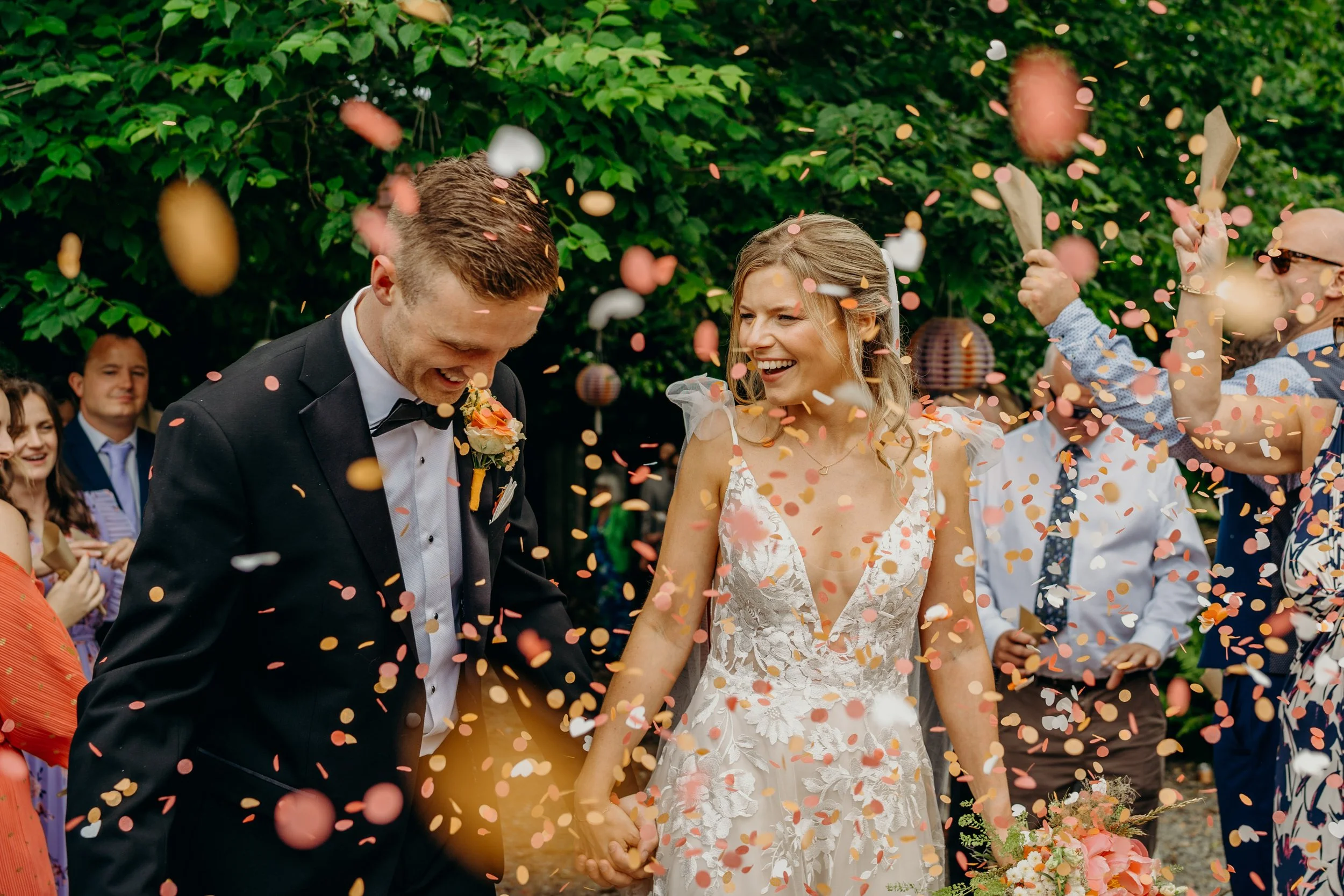 A bride and groom holding hands and smiling as they are showered with pink and white confetti at their wedding celebration outdoors, with friends in the background.