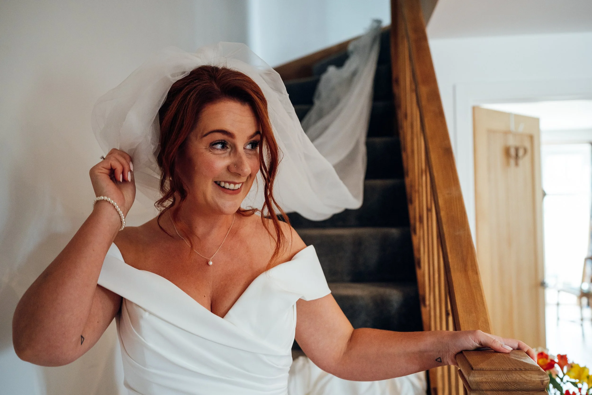 A woman with red hair in a white wedding dress and veil smiling on a staircase.