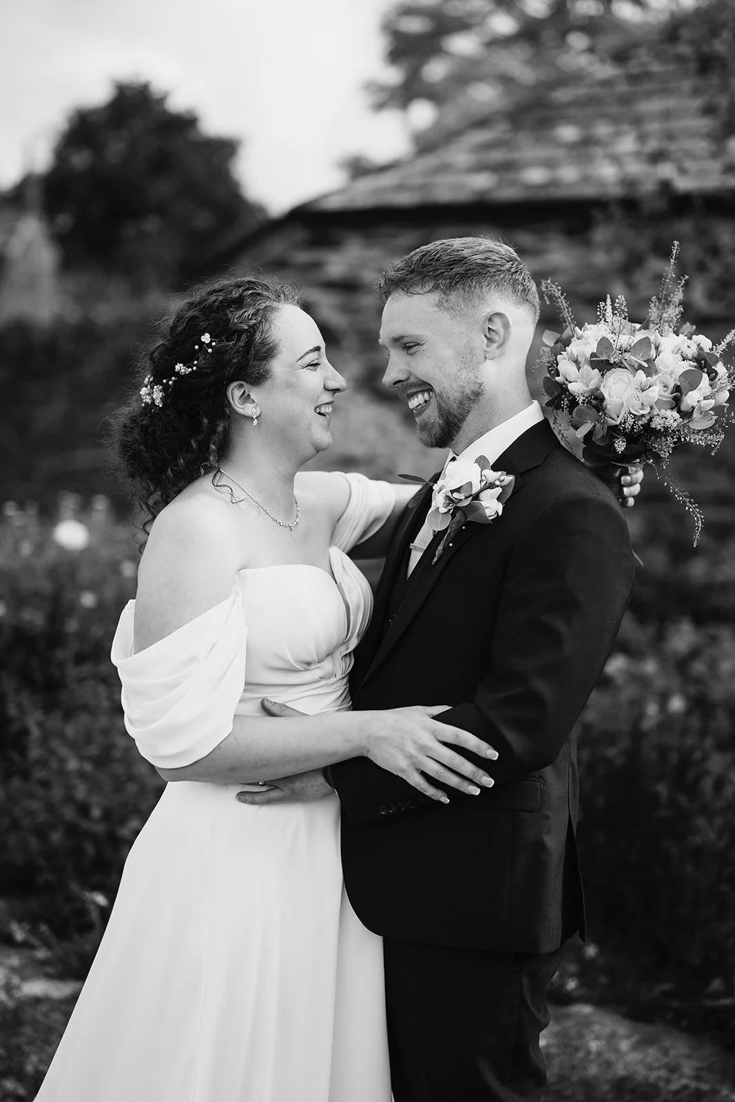 Black and white photo of a bride and groom smiling and holding each other on their wedding day. The bride has curly hair decorated with small flowers and is wearing an off-the-shoulder dress, while the groom has a beard, mustache, and is dressed in a