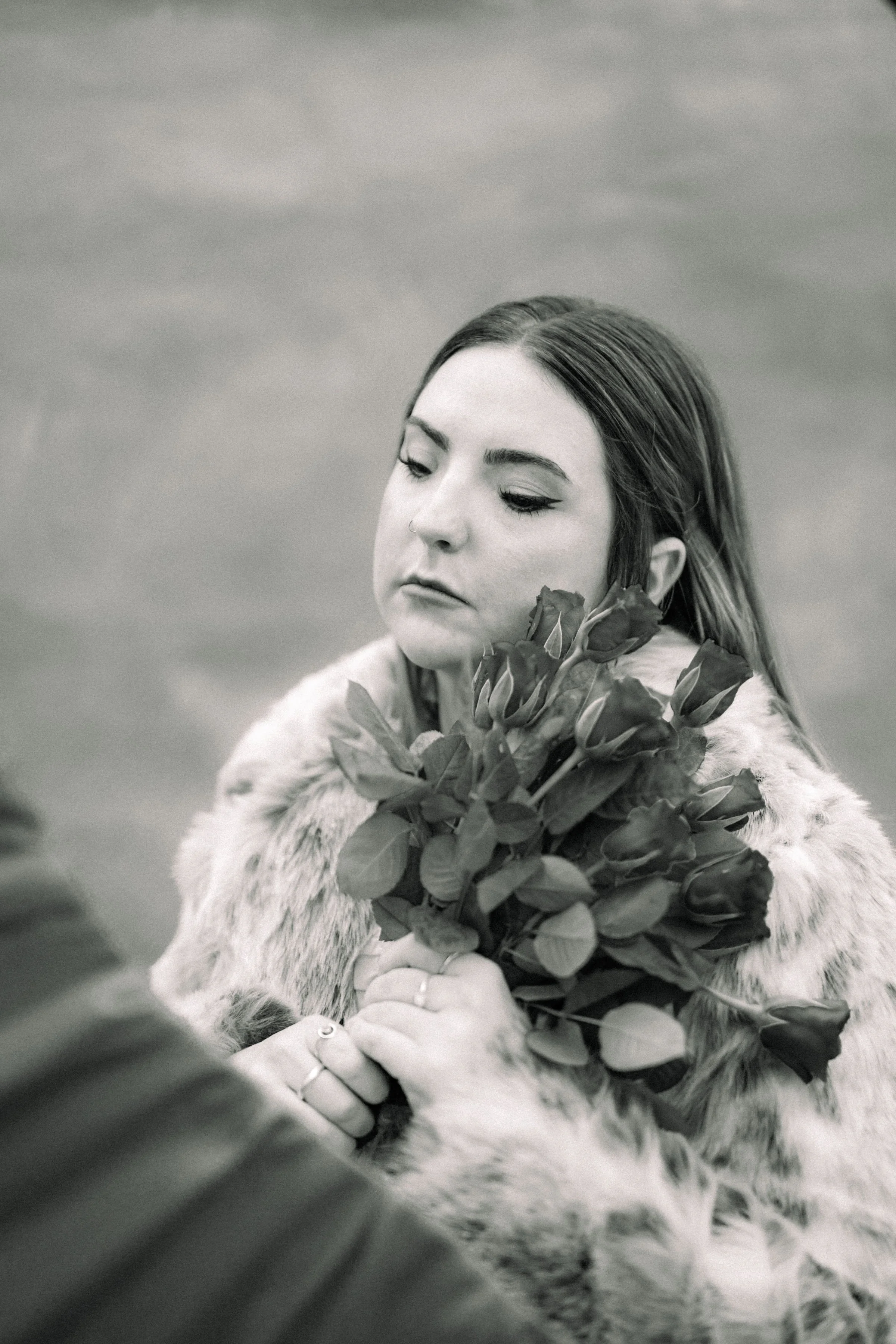 A woman with long hair, wearing a fur coat, holds a bouquet of roses and looks down thoughtfully.