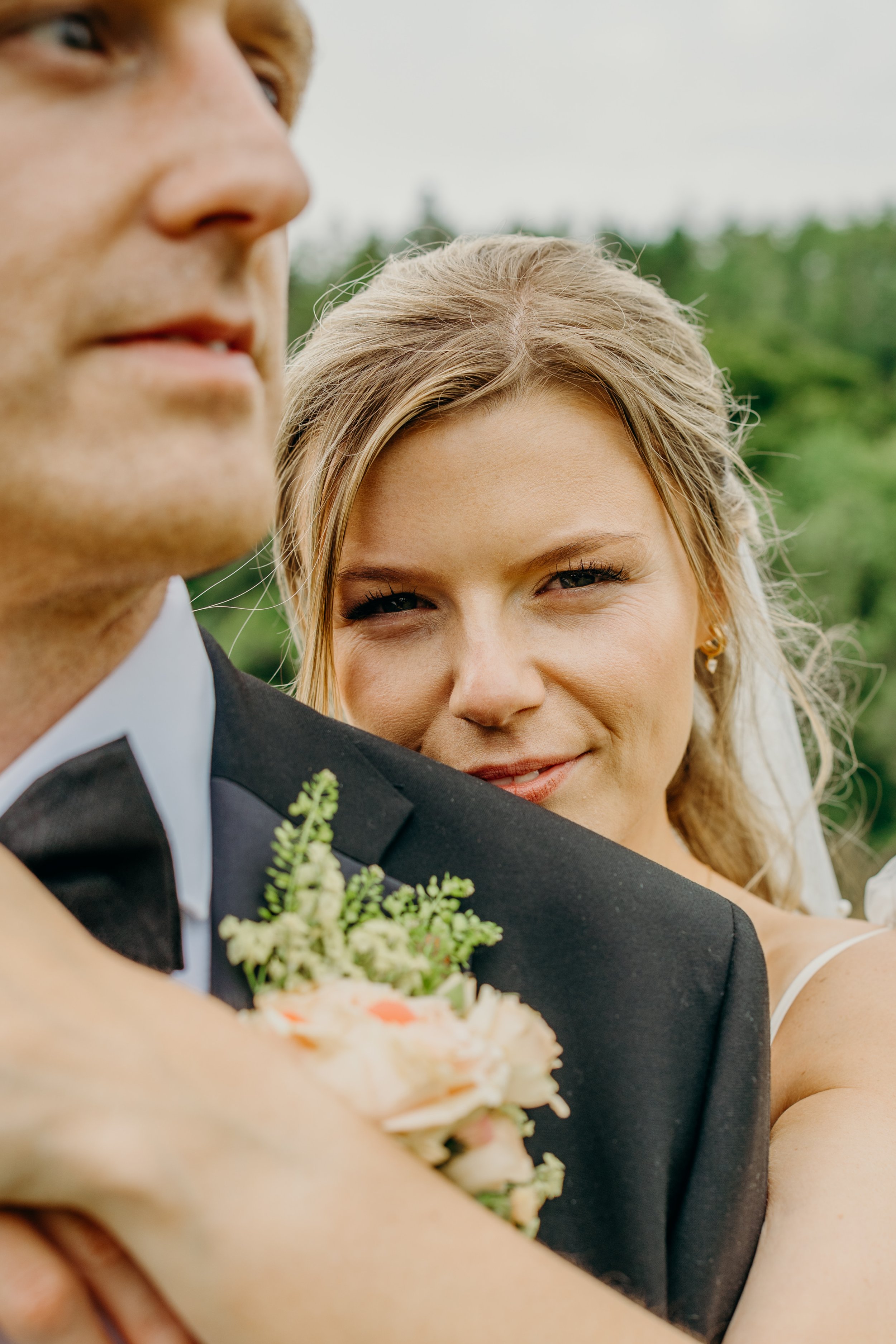 A bride hugging a groom from behind, smiling at the camera outdoors with green trees in the background.