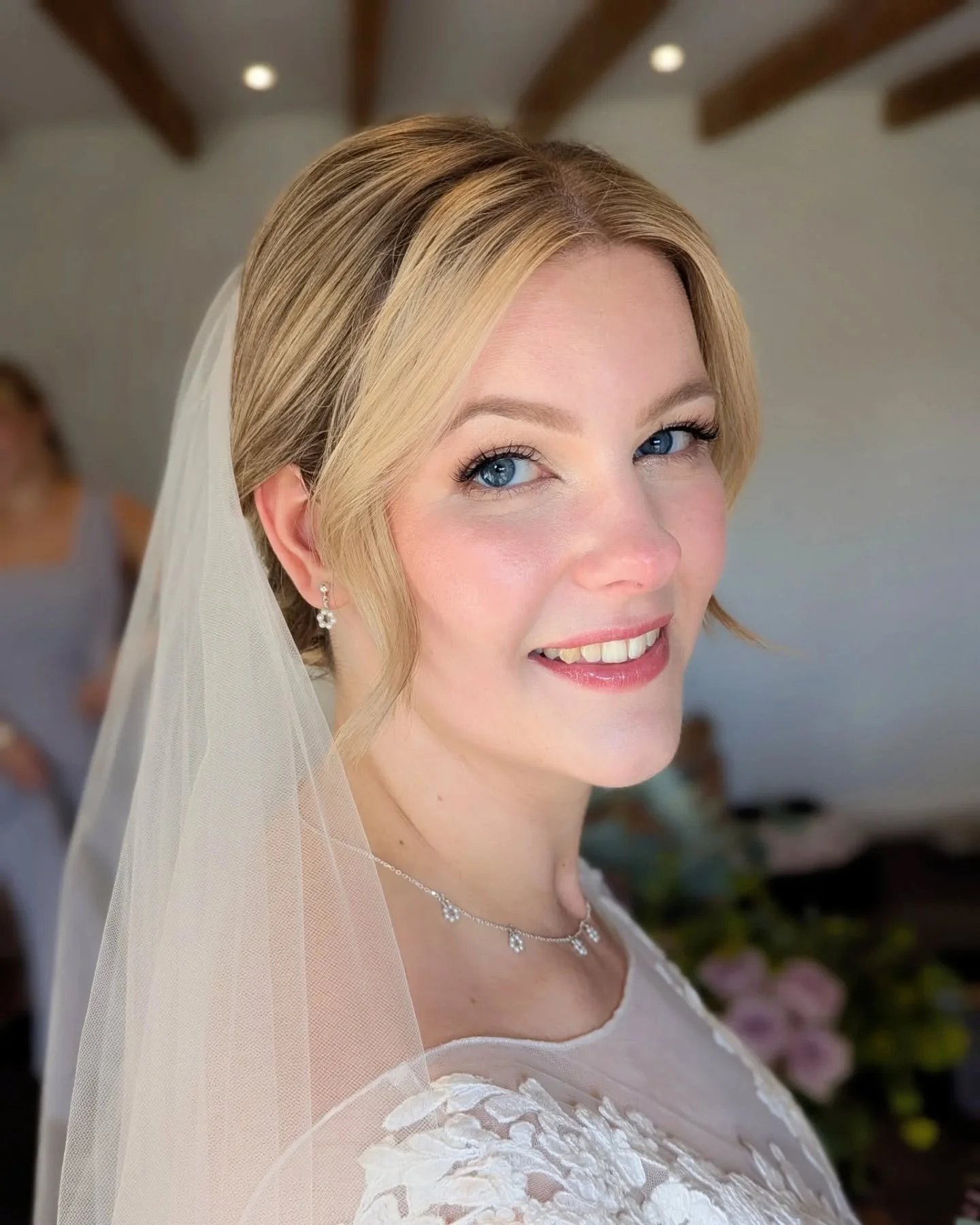 Close-up of a smiling bride with blonde hair, blue eyes, wearing a white wedding dress and jewelry, with a veil and a blurred background showing indoor setting.
