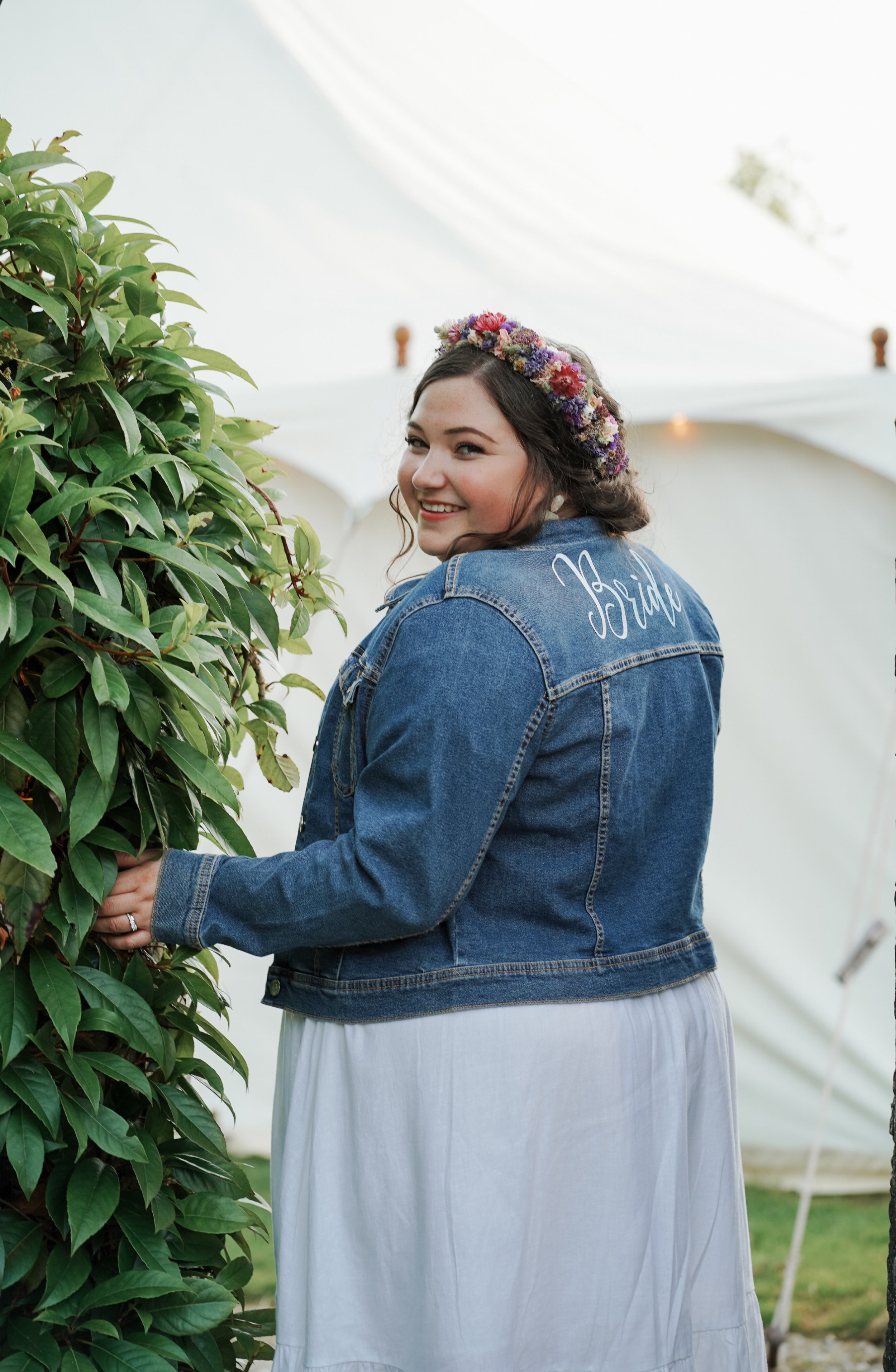 A smiling bride in a denim jacket with 'Bride' written on the back, standing outdoors near green bushes, wearing a floral crown, and holding a branch.