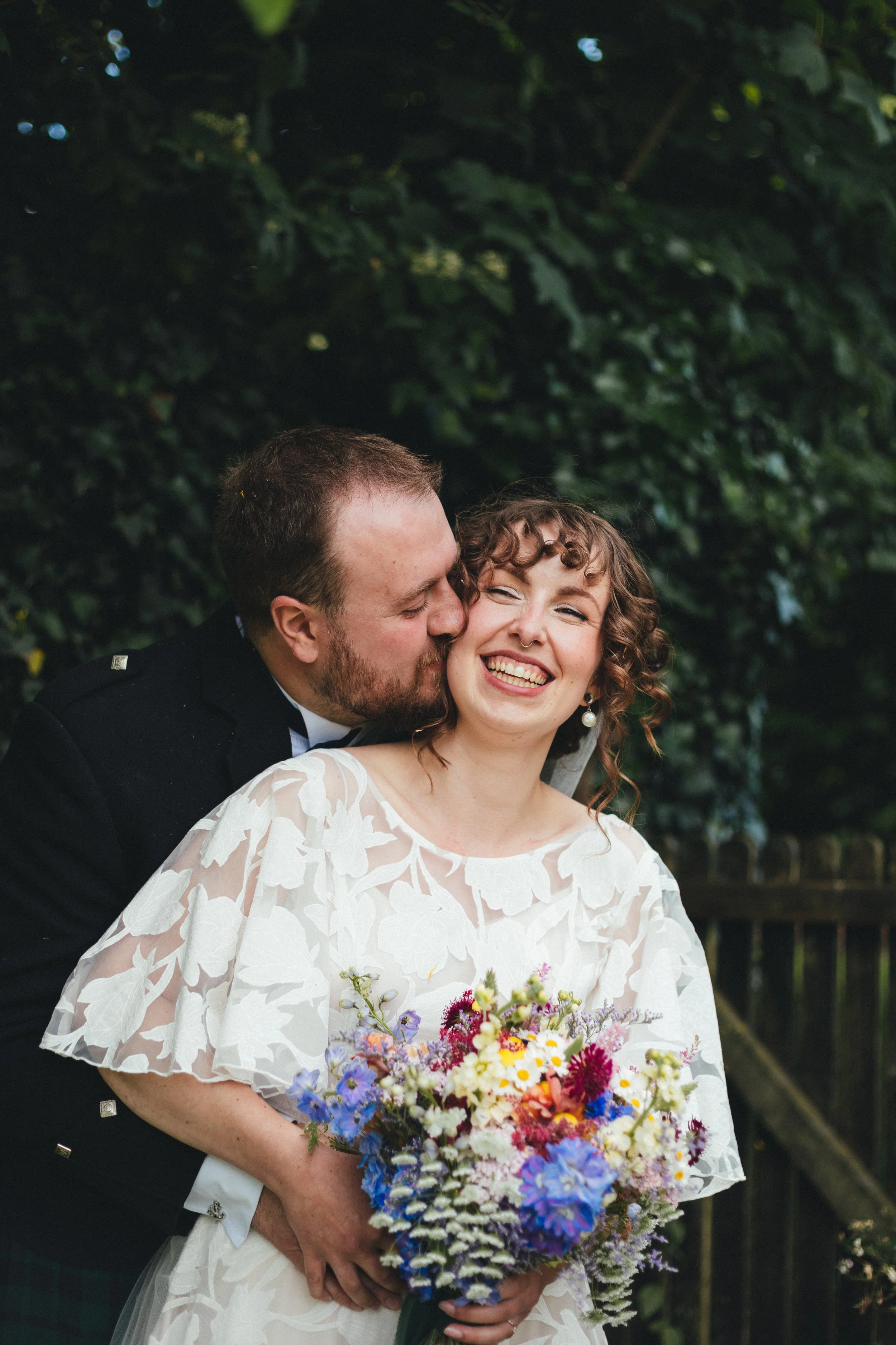 A man and a woman at a wedding, with the man kissing the woman's cheek, the woman smiling, holding a large bouquet of flowers. They stand outdoors in front of a leafy background.