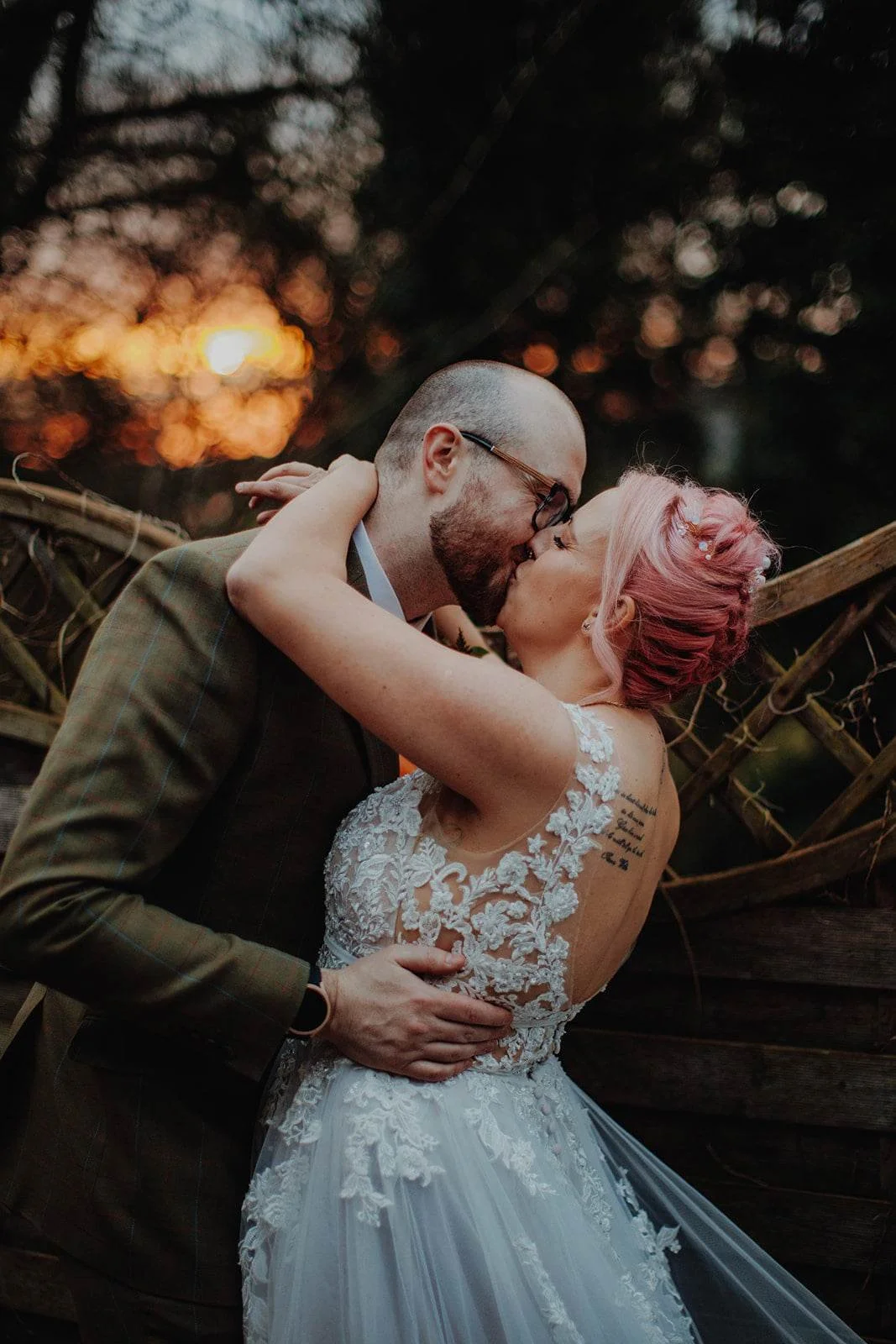 A couple sharing a kiss at sunset, the woman with pink hair wearing a white lace wedding dress, and the man with glasses in a brown suit, in an outdoor setting with trees and a wooden fence.