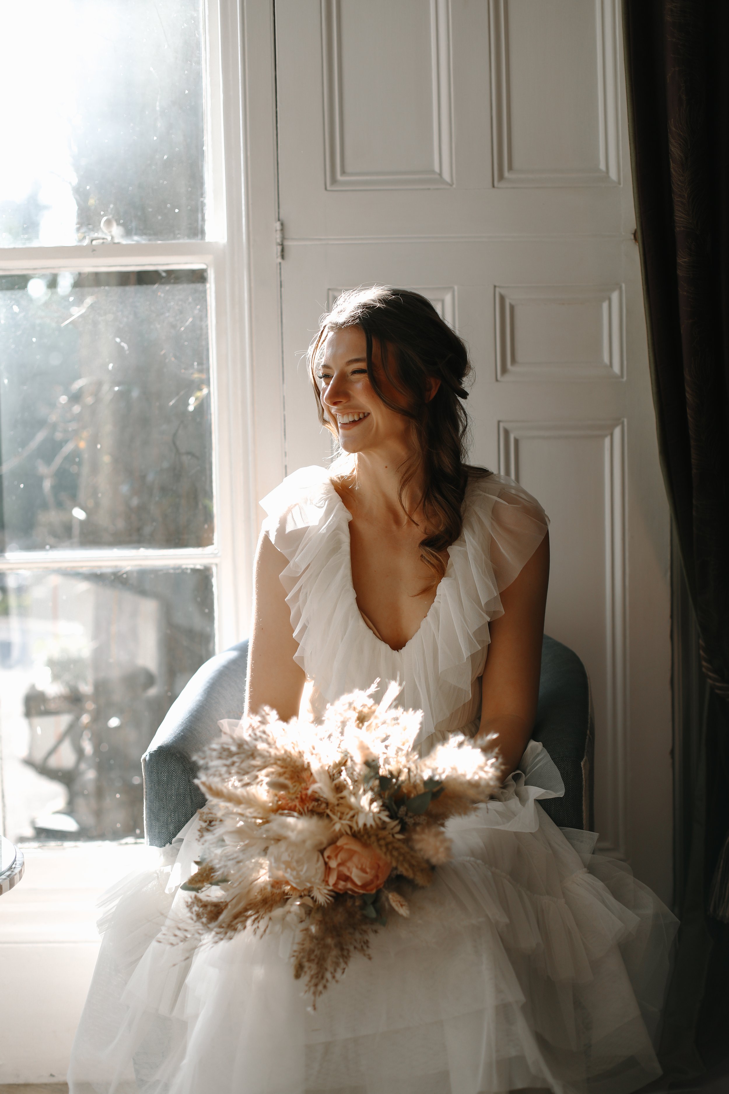 A smiling woman in a wedding dress sitting by a window, holding a bouquet of flowers.