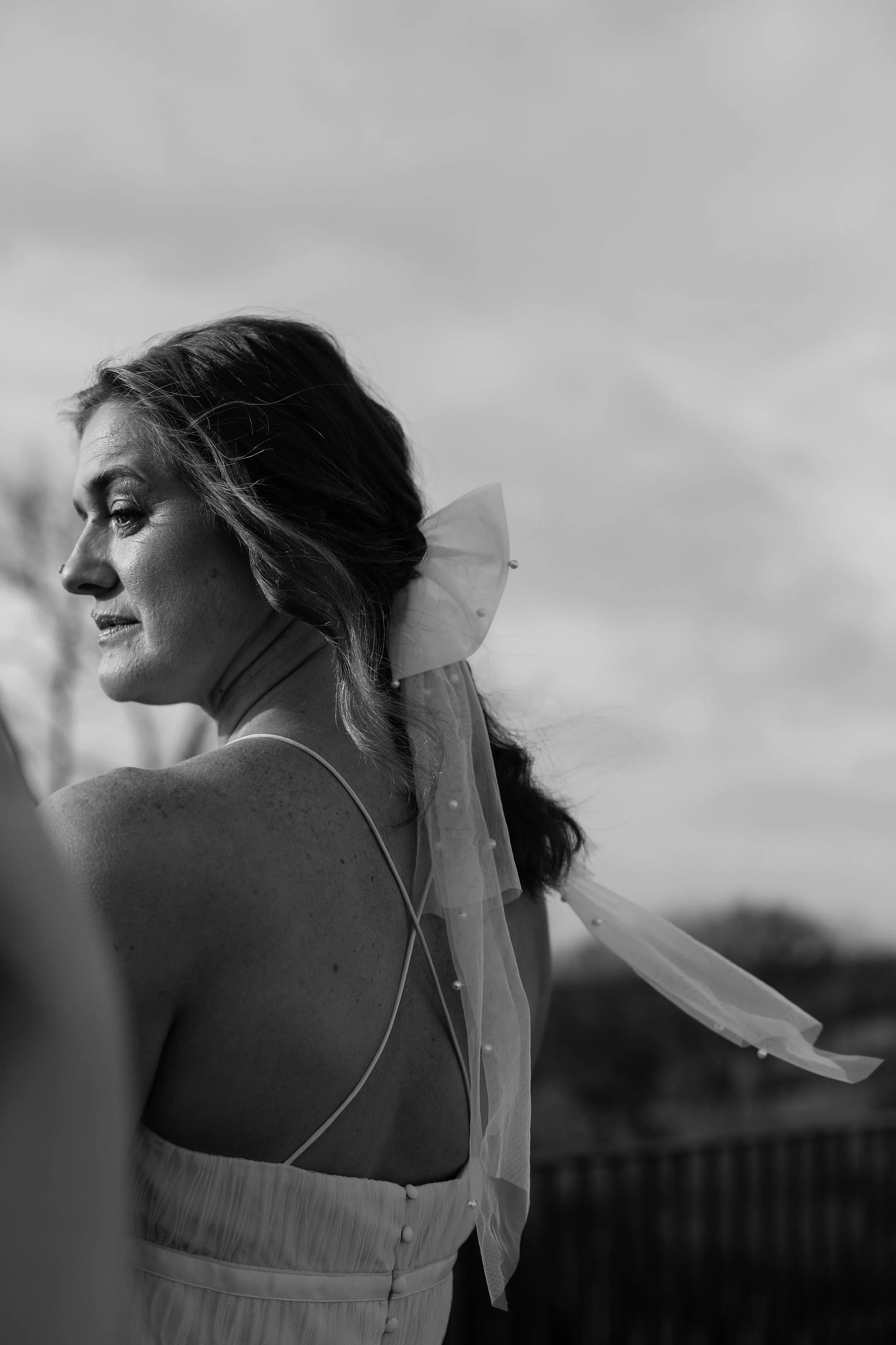 Black and white photo of a woman with wavy hair, looking downward, wearing a spaghetti strap dress with a sheer ribbon tied into a bow on her hair.