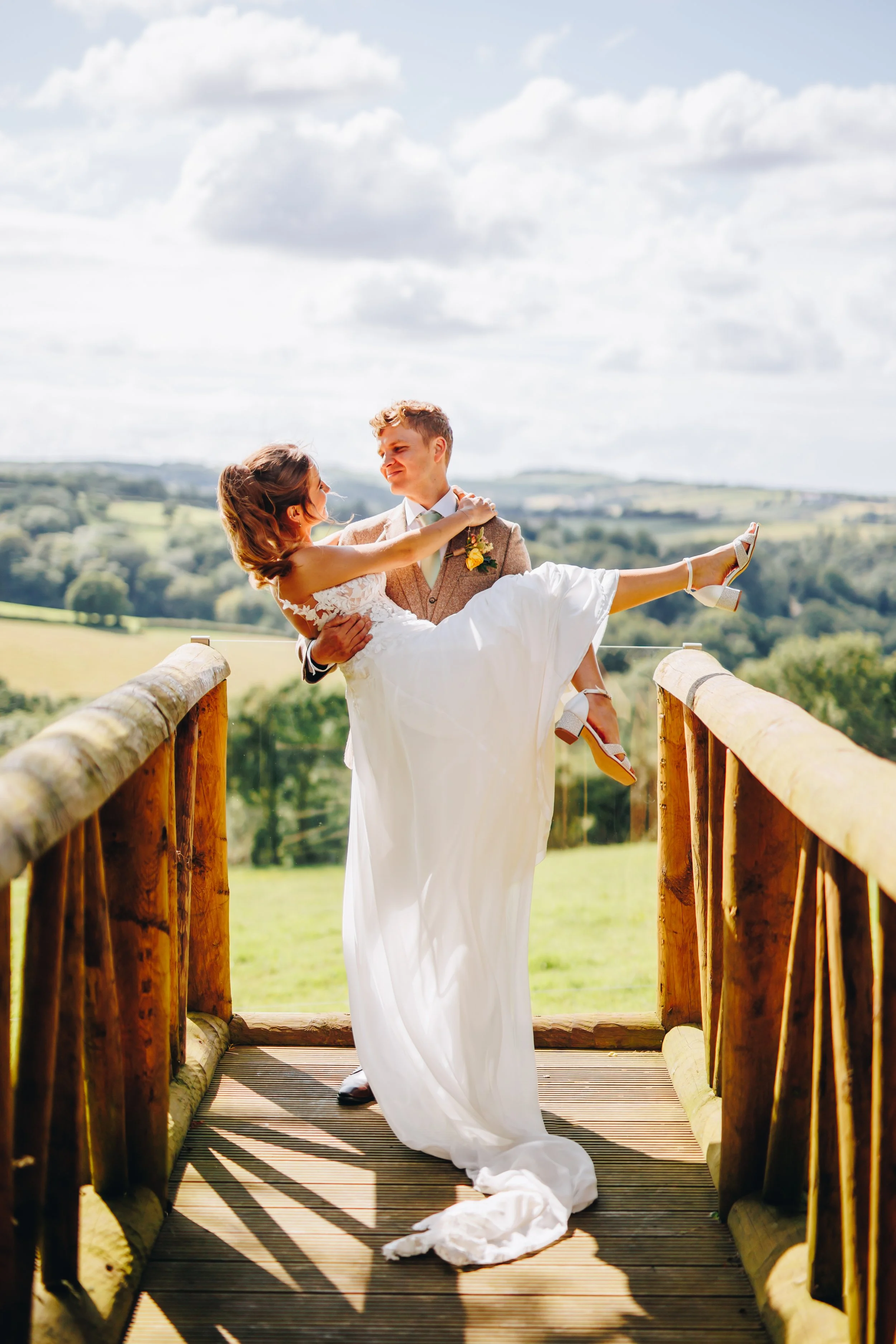 A groom carrying a bride, who is wearing a wedding dress, on a wooden bridge outdoors with a scenic green landscape and cloudy sky in the background.