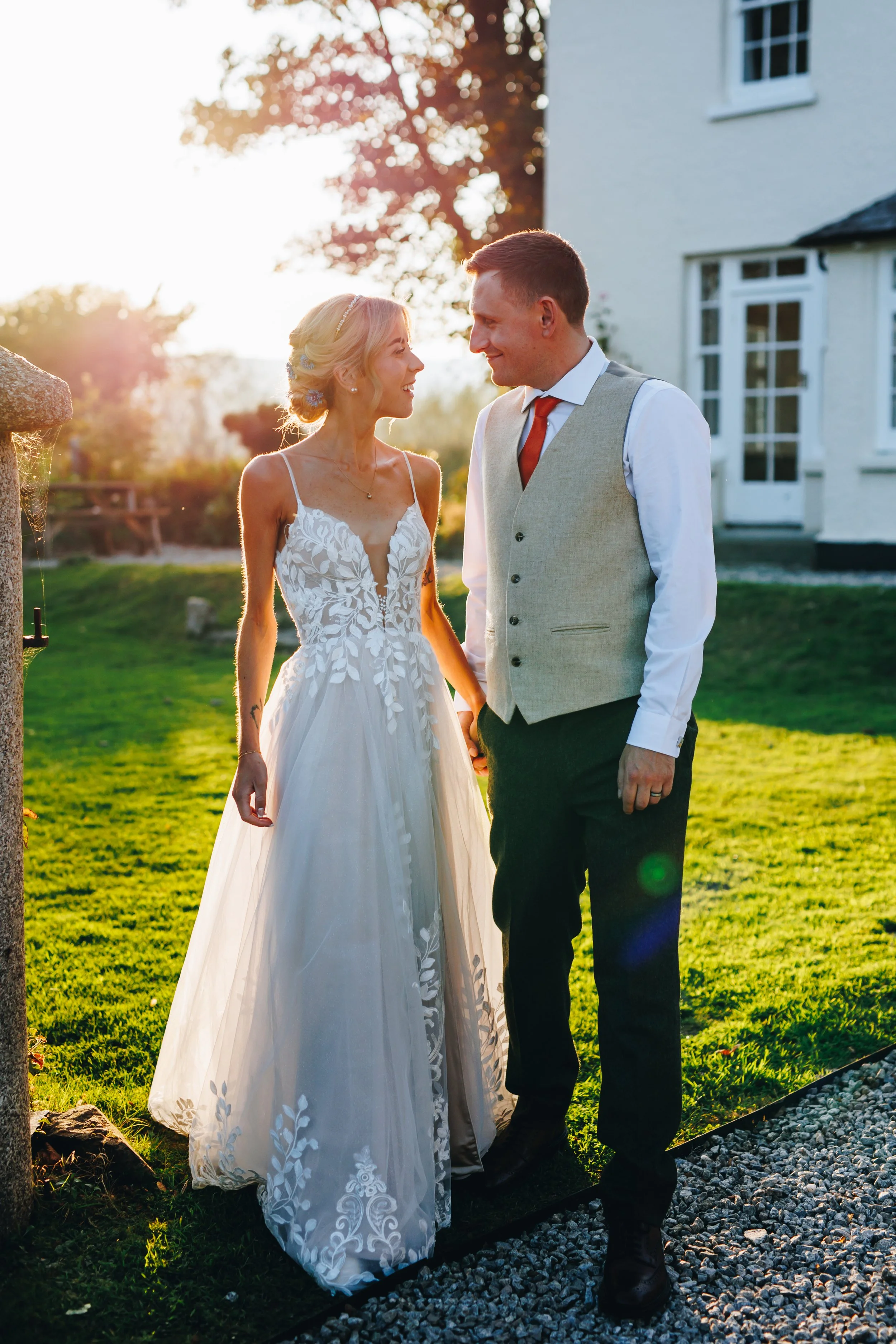 A bride and groom standing outdoors during sunset, gazing at each other, holding hands, with a house and garden in the background.