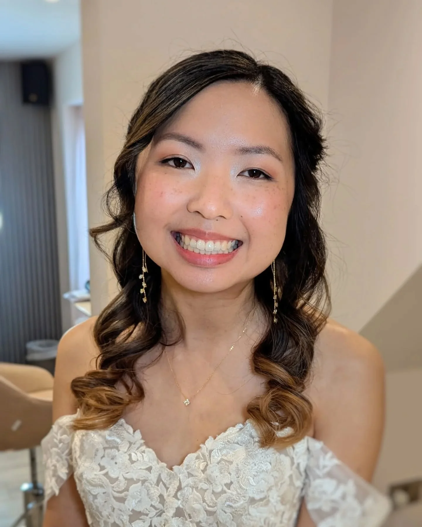 A smiling woman with wavy hair, wearing a white lace dress, gold jewelry, and makeup, in a well-lit indoor space.