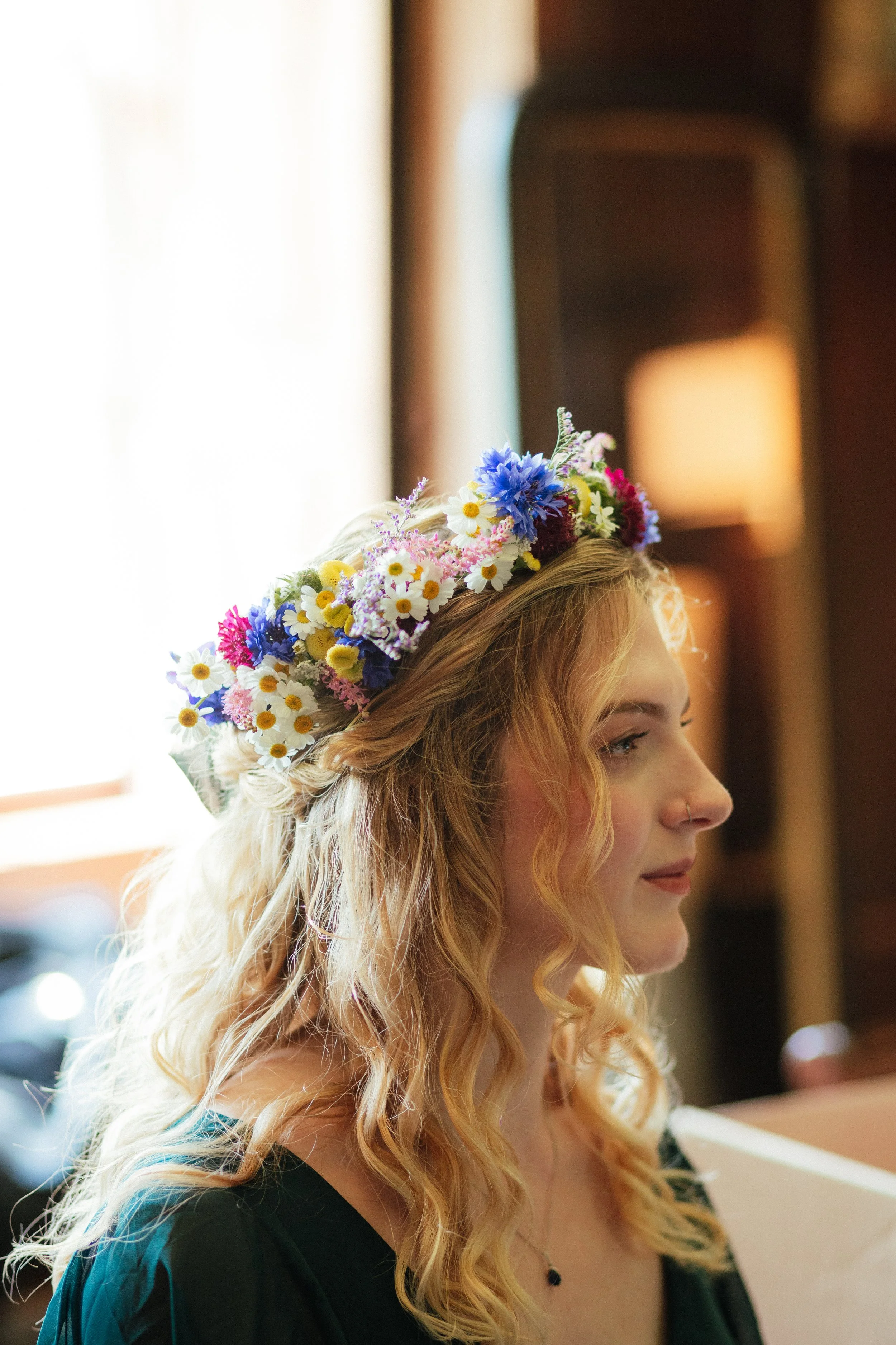 A young woman with curly blonde hair and a nose piercing wearing a floral crown made of daisies, cornflowers, and other colorful flowers, looking to the side indoors with sunlight streaming through a window.