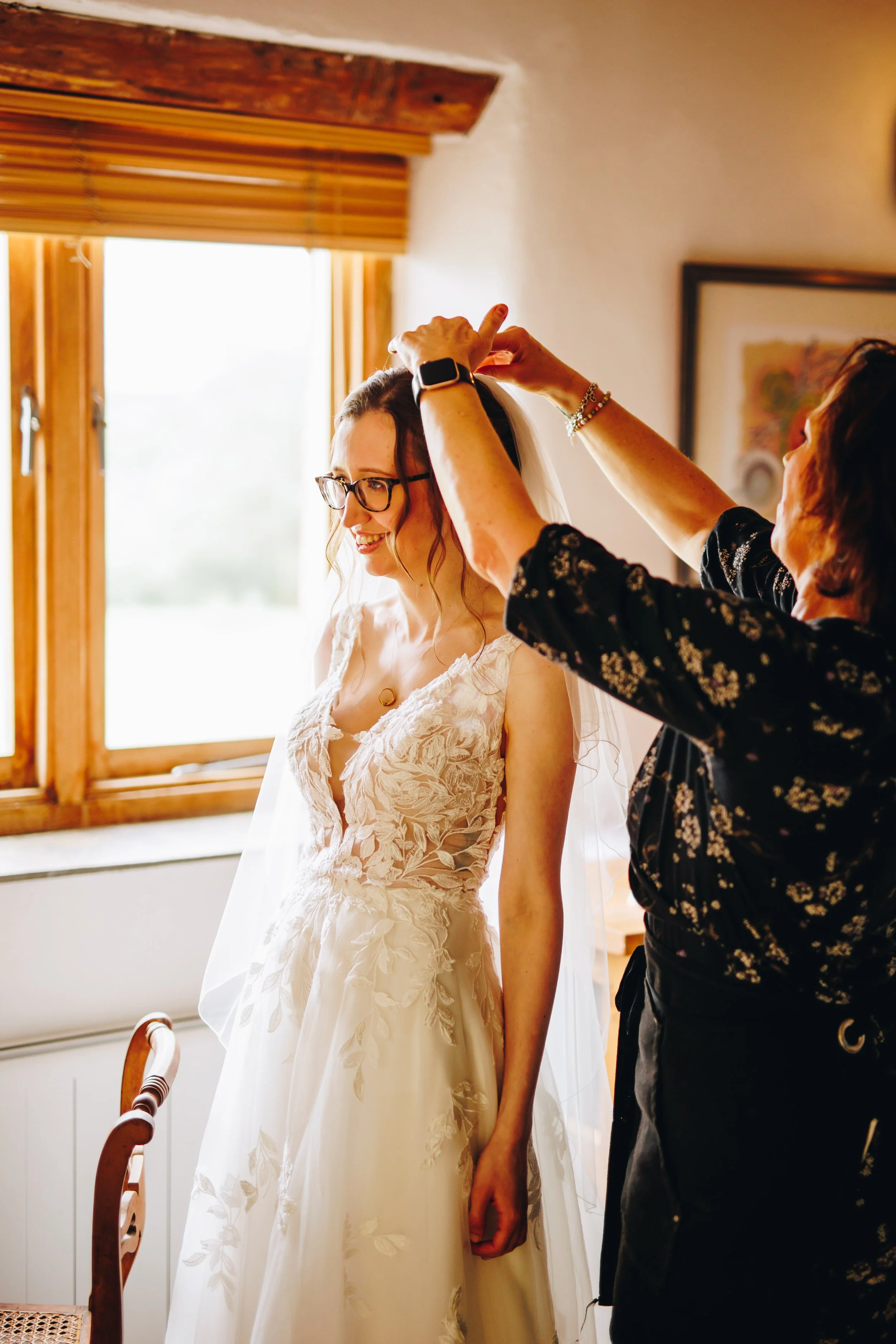 A bride with glasses wearing a lace wedding gown, smiling as someone helps her adjust her veil in a warmly lit room with wooden window frames.