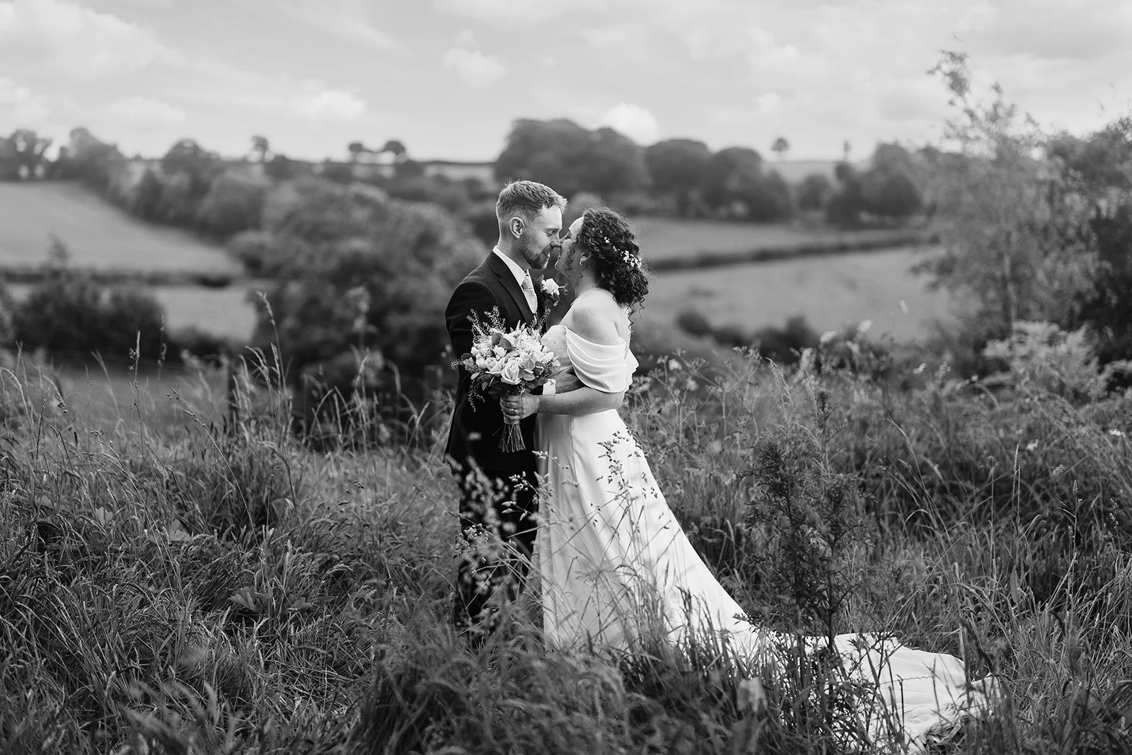 A black-and-white photo of a bride and groom standing in a field, close to each other, holding a bouquet of flowers, with rolling hills and trees in the background, during a wedding photoshoot.