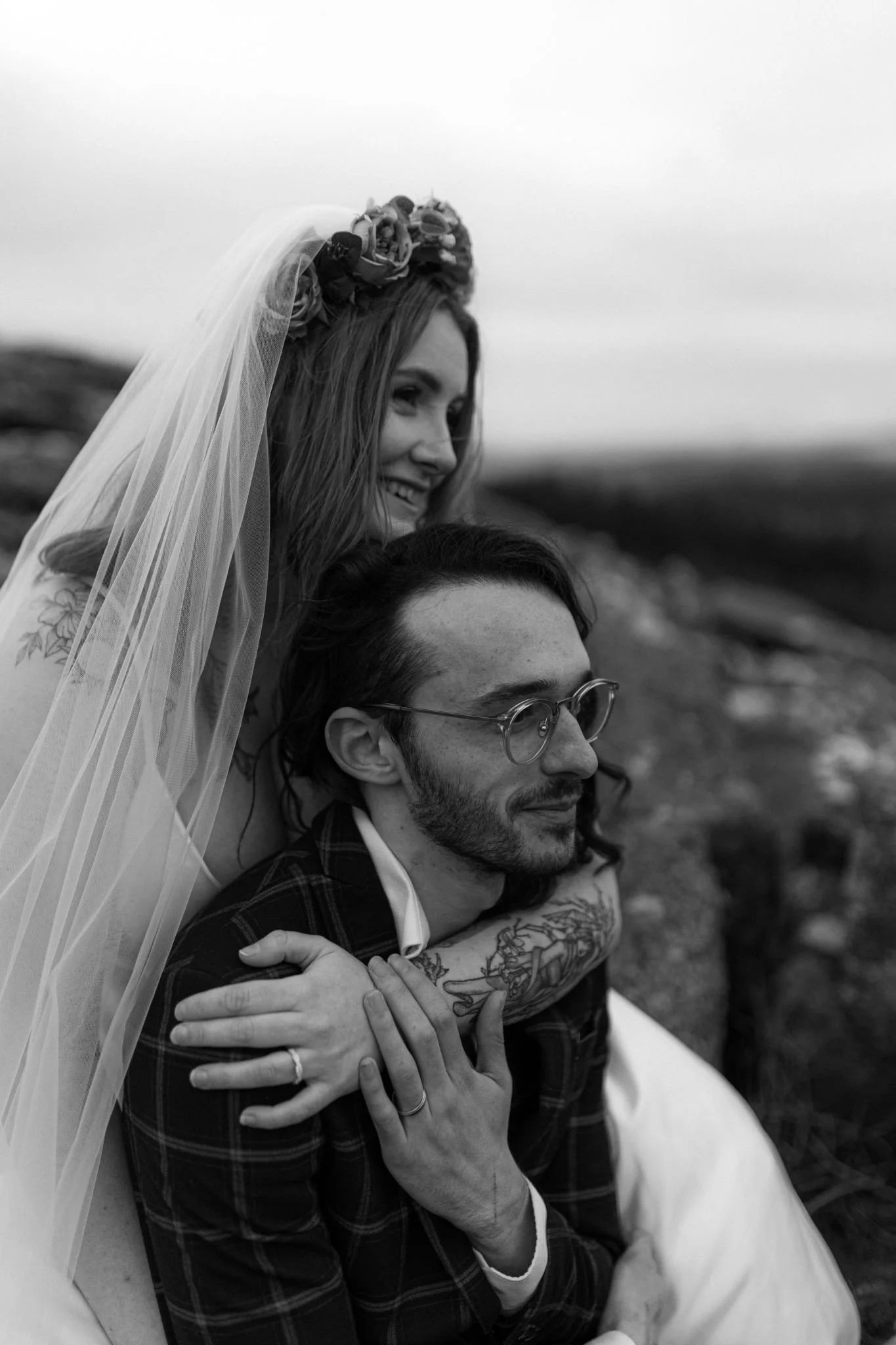 A black and white photo of a woman in a wedding dress and veil, embracing a man with glasses and tattoos, sitting outdoors on a rocky landscape.
