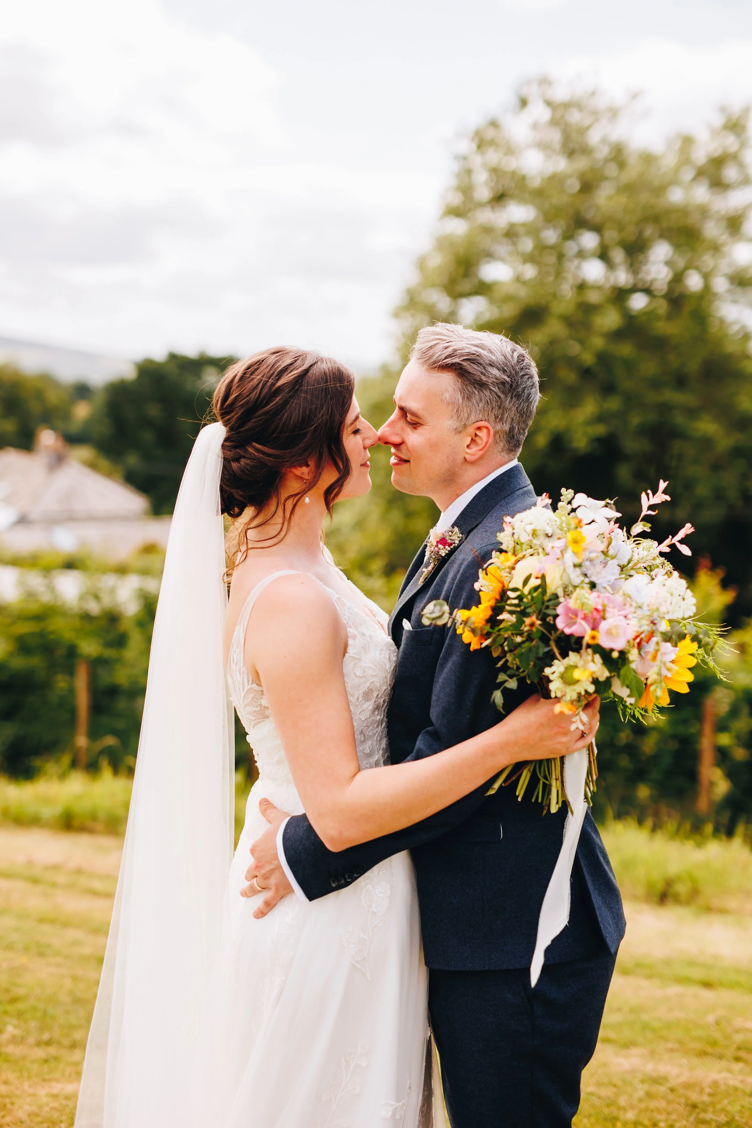 Bride and groom embrace outdoors during wedding, holding a bouquet of colorful flowers, with trees and cloudy sky in the background.