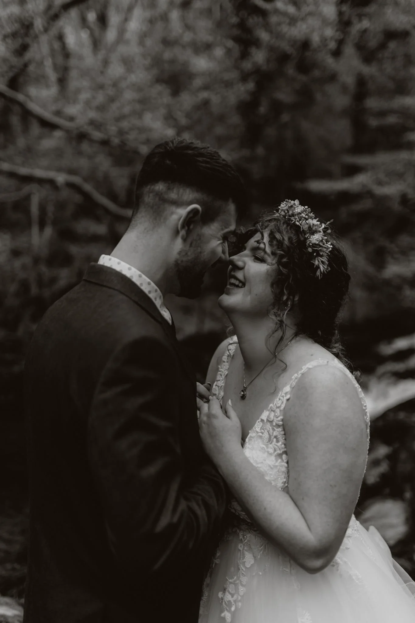 A black-and-white photograph of a bride and groom happily embracing outdoors, with the bride wearing a lace wedding dress and floral crown and the groom in a suit.