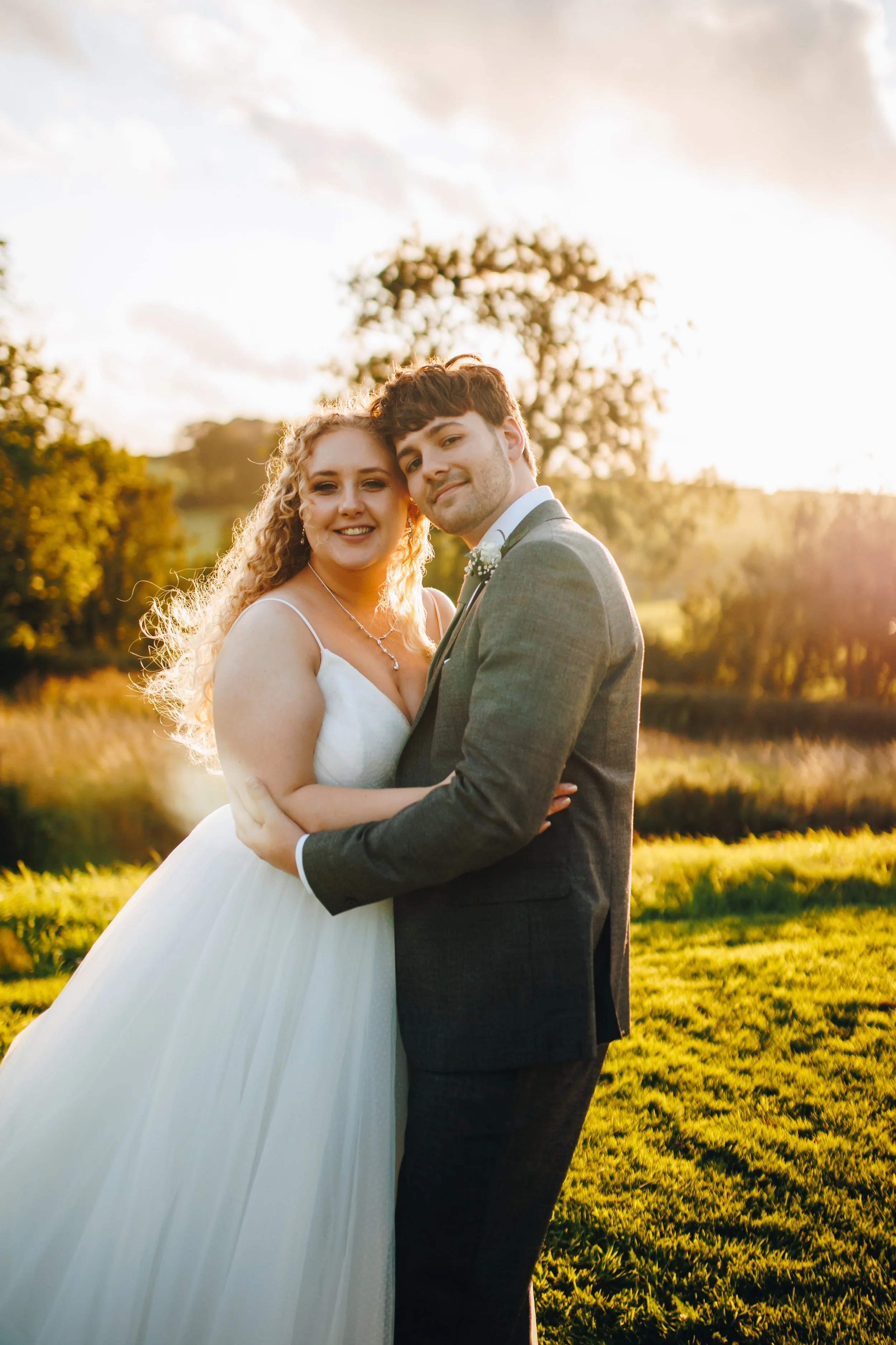 A bride and groom smiling and hugging outdoors at sunset on their wedding day.