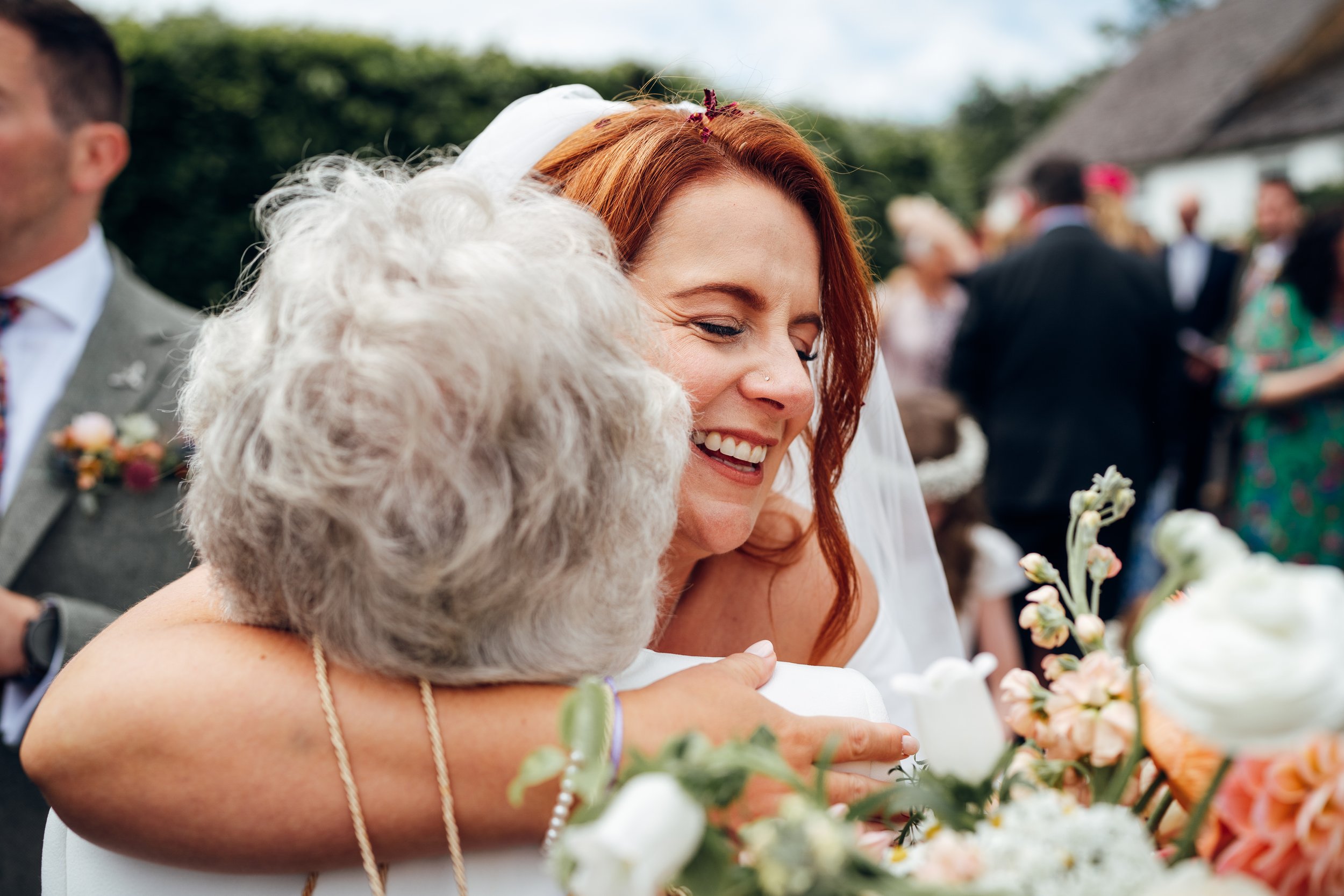 A woman with red hair and a white dress hugging an elderly woman with gray hair during a wedding celebration, with other guests in the background.