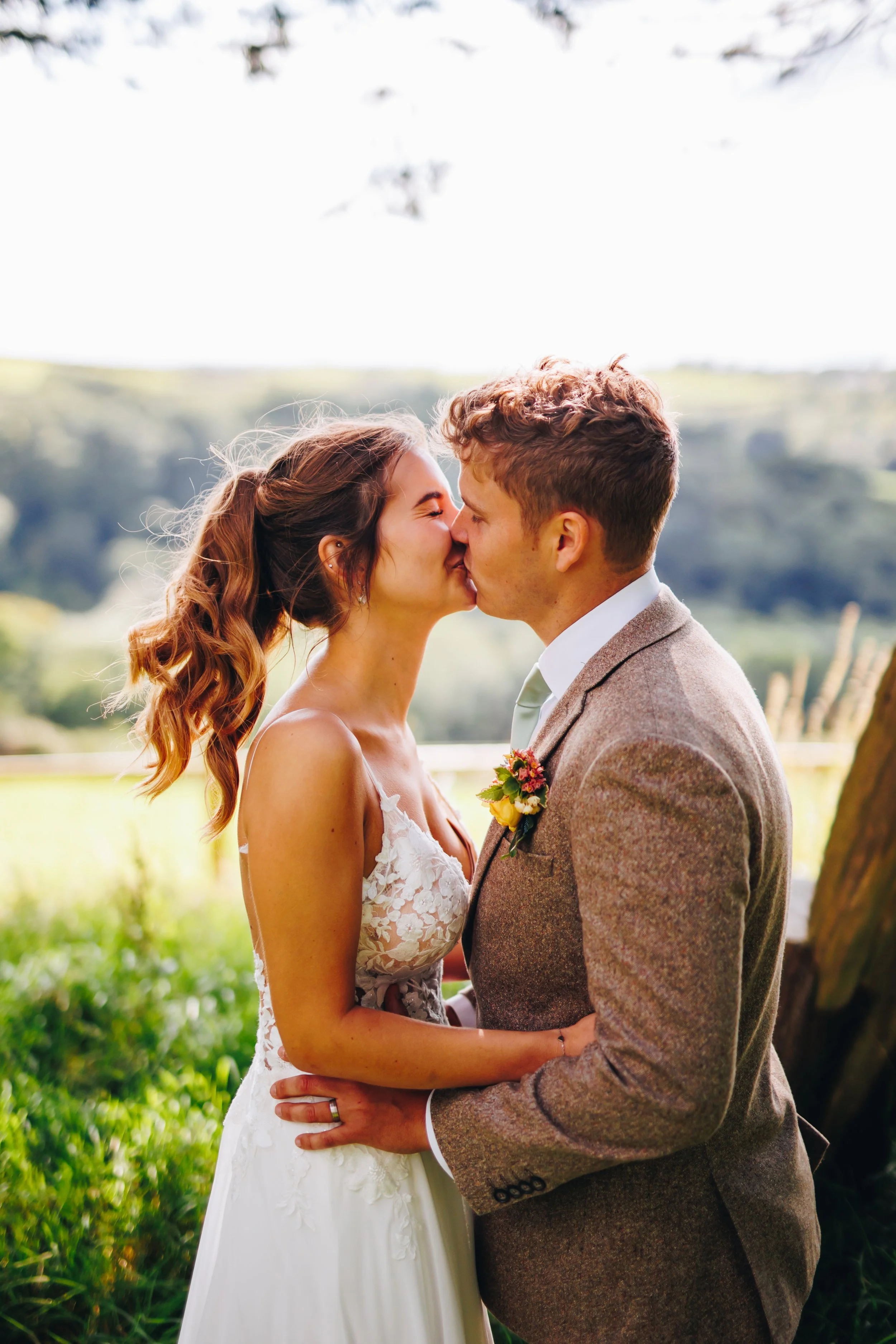 A bride and groom kiss outdoors, both dressed in wedding attire, with a scenic landscape of green hills and trees in the background.