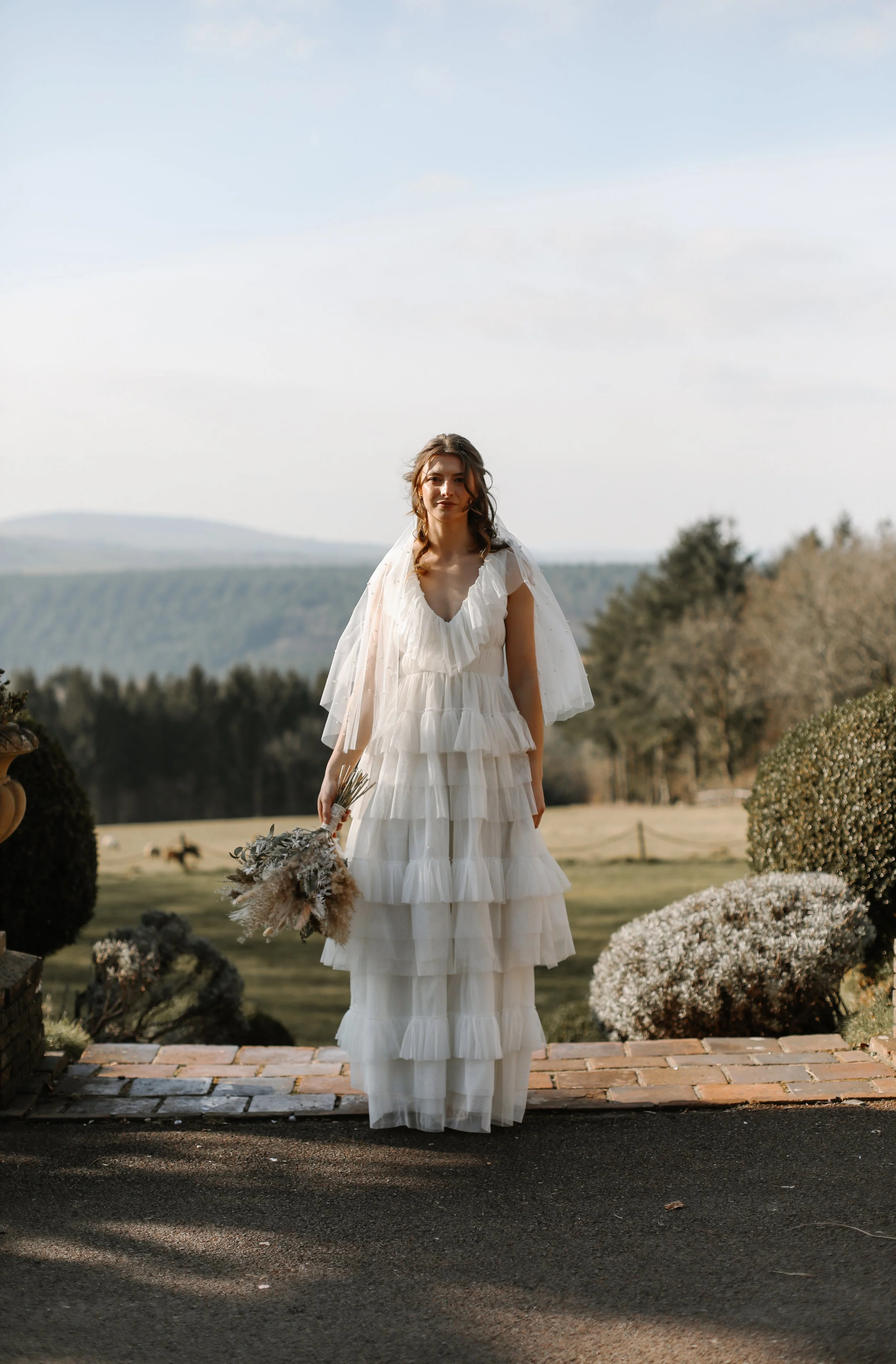 A woman in a white, tiered, ruffled dress holding a bouquet of dried flowers, standing outdoors on a brick path with a scenic background of trees and rolling hills under a cloudy sky.