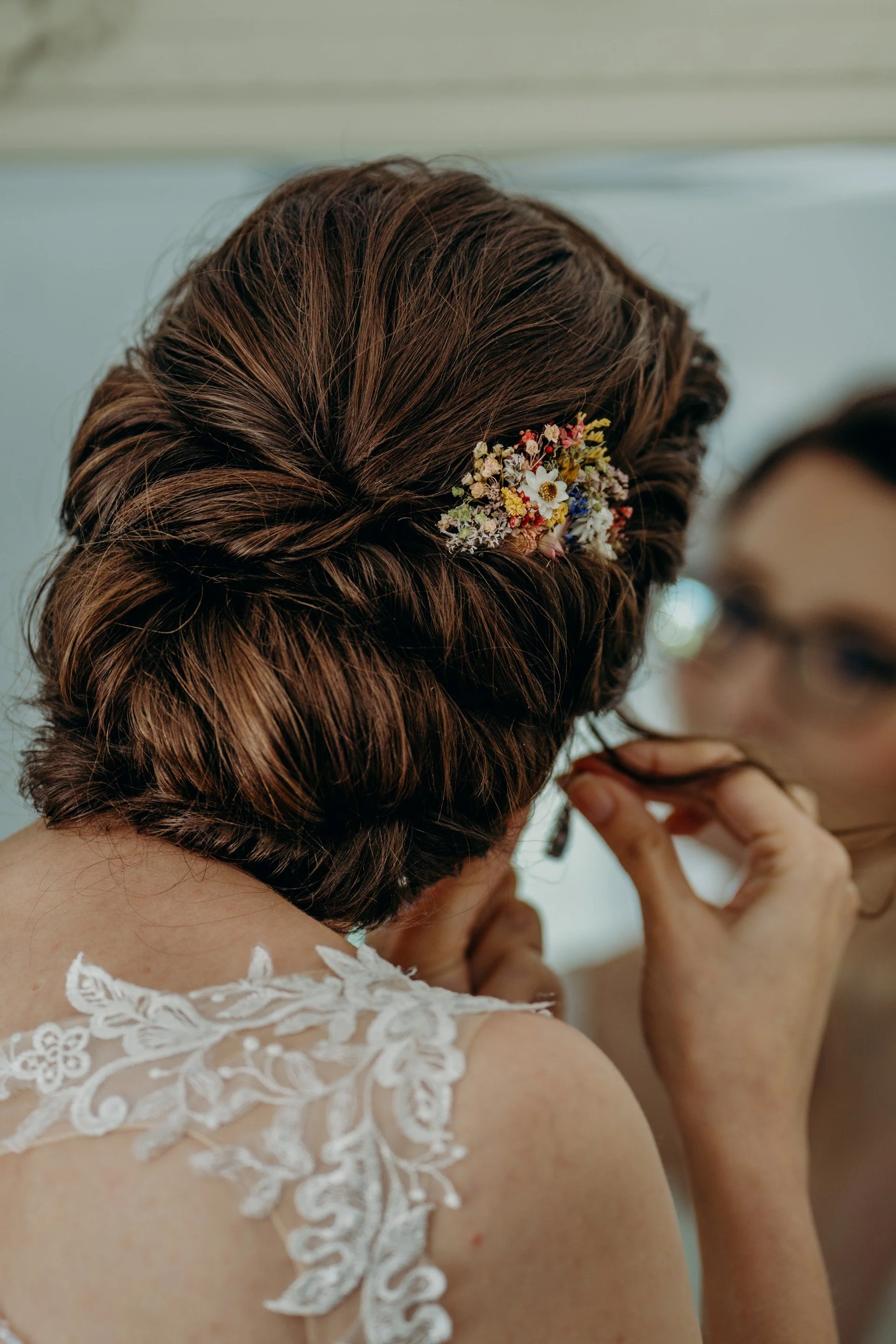 Close-up of a woman with styled brown hair wearing a floral hairpiece, preparing her earring, with a mirror reflection of her face with glasses.