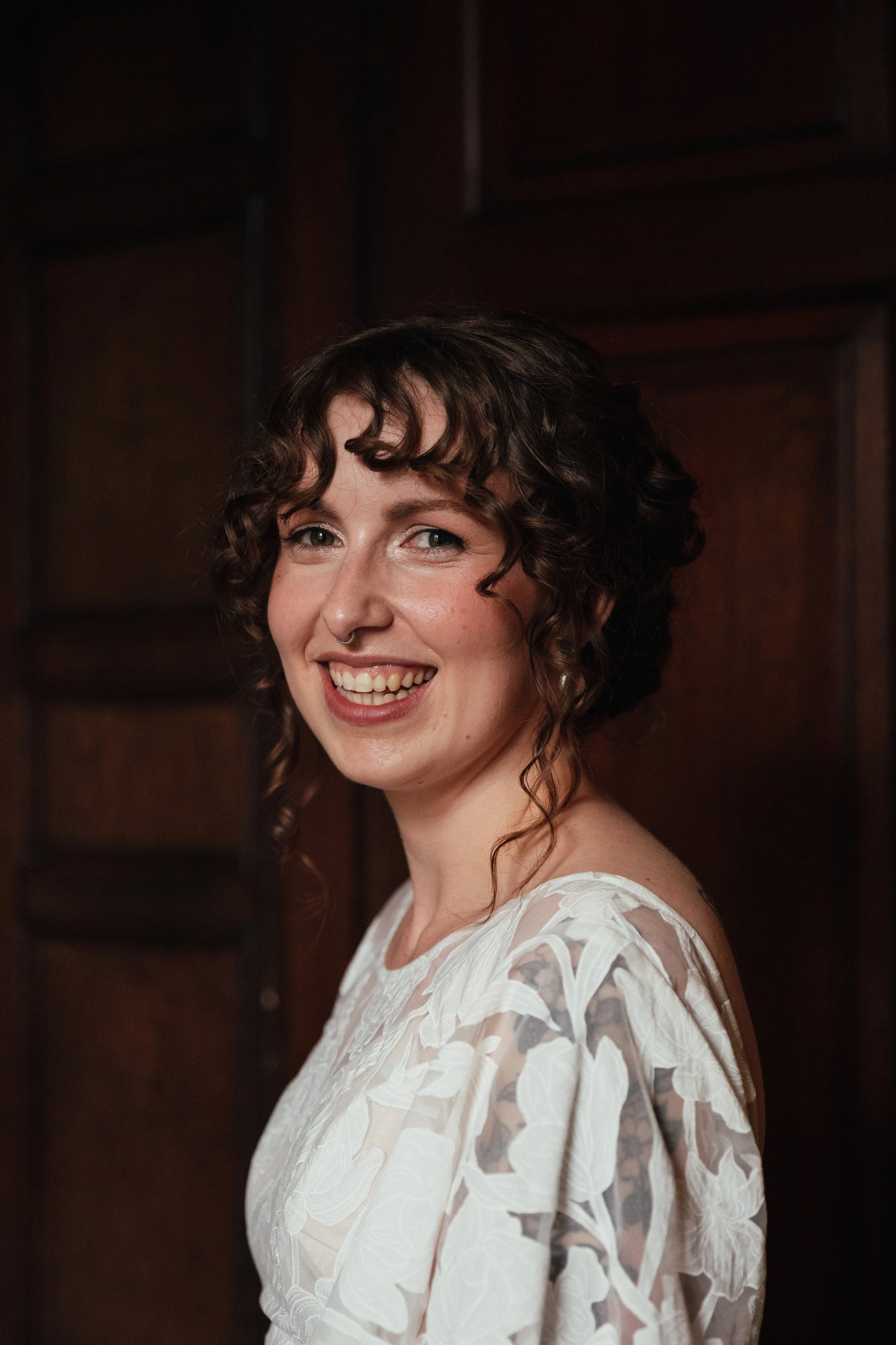 A smiling woman with curly brown hair, fair skin, wearing a white floral lace dress, standing in front of a dark wooden background.