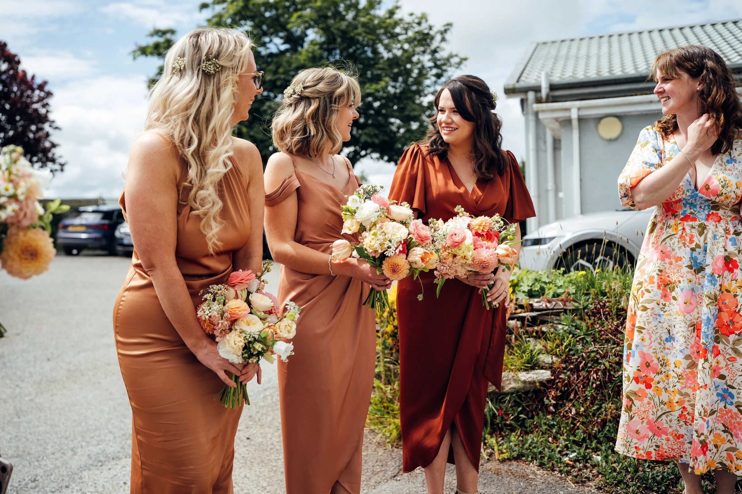 Four women in floral dresses holding bouquets, chatting outdoors near a house with cars parked in background.