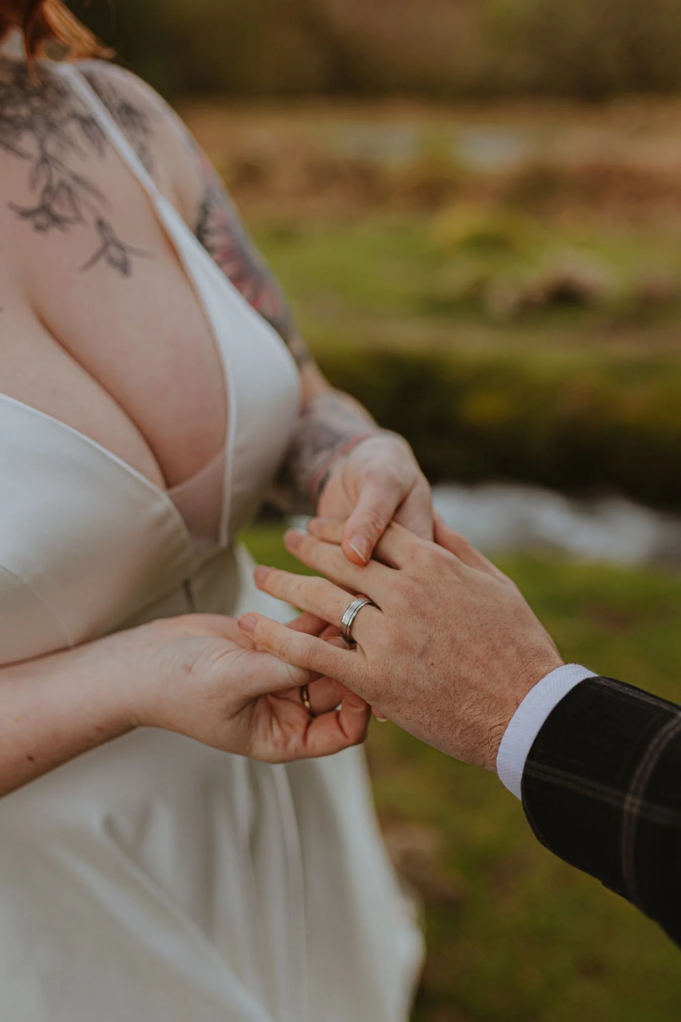 Close-up of a couple exchanging wedding rings outdoors, with a blurred natural background. The woman is wearing a white satin dress with floral tattoos on her shoulder, and the man is wearing a dark patterned suit.