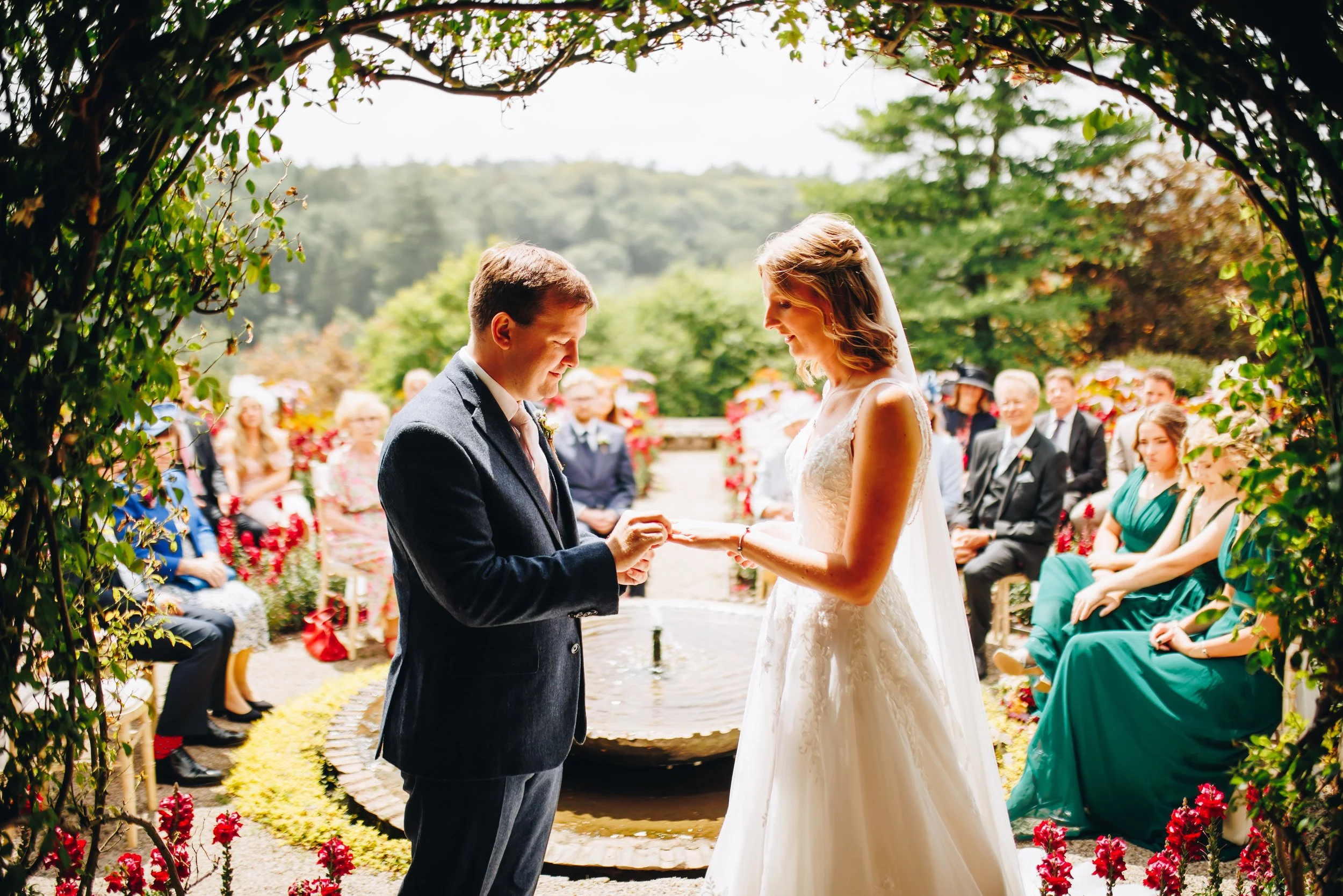 A couple exchanging rings during an outdoor wedding ceremony, surrounded by guests seated on either side, with a lush green landscape in the background.