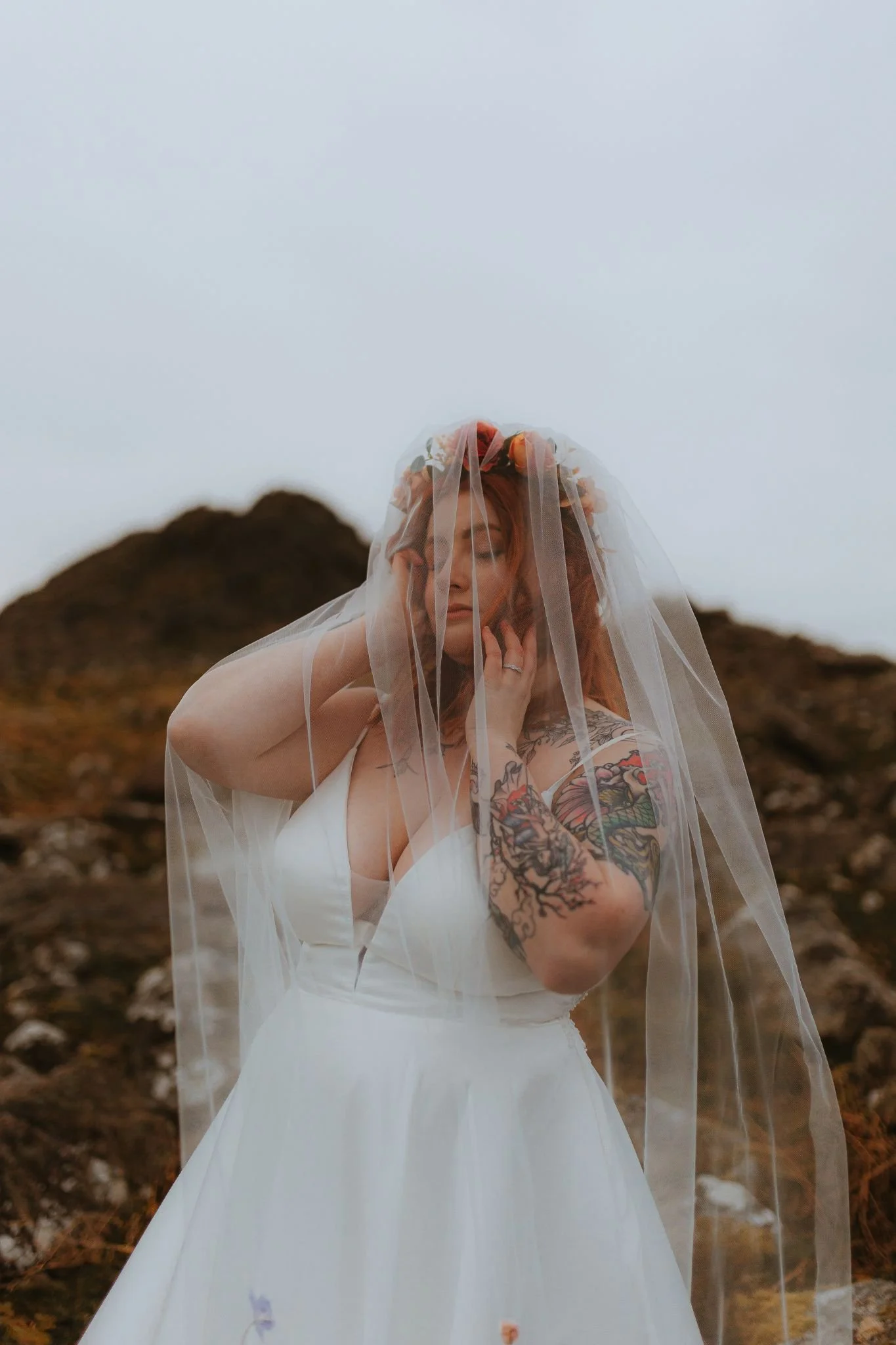 A woman in a white wedding dress with tattoos on her arm and a floral headband, standing outdoors under a cloudy sky with mountains in the background, wearing a wedding veil.