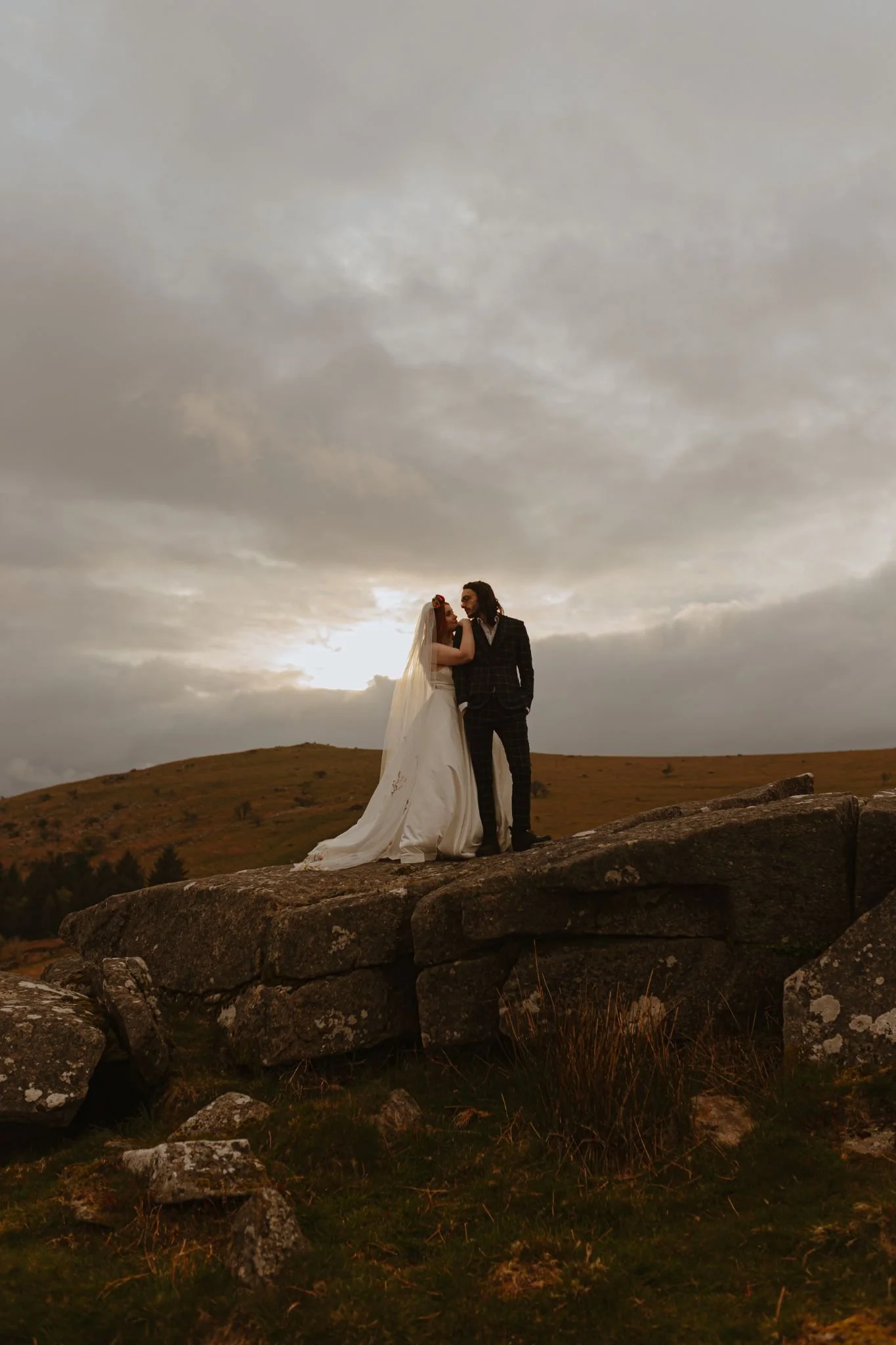 A bride and groom standing on a large rock formation outdoors during sunset, with a cloudy sky overhead.
