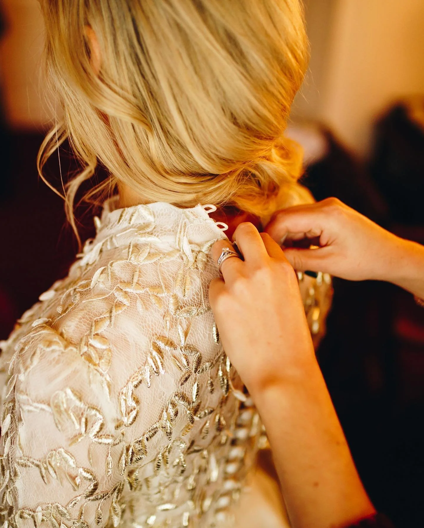 Close-up of a woman with blonde hair, being assisted with jewelry or a dress, wearing a white lace dress with metallic embroidery.