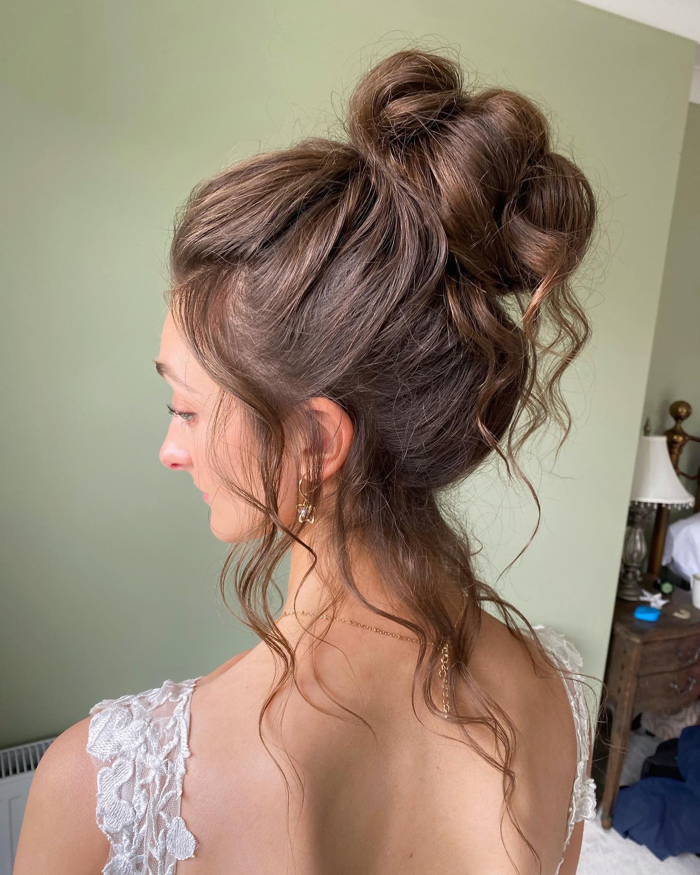 A woman with styled, curly brown hair in an elaborate updo, wearing earrings and a lace dress, with a green wall and a wooden dresser in the background.