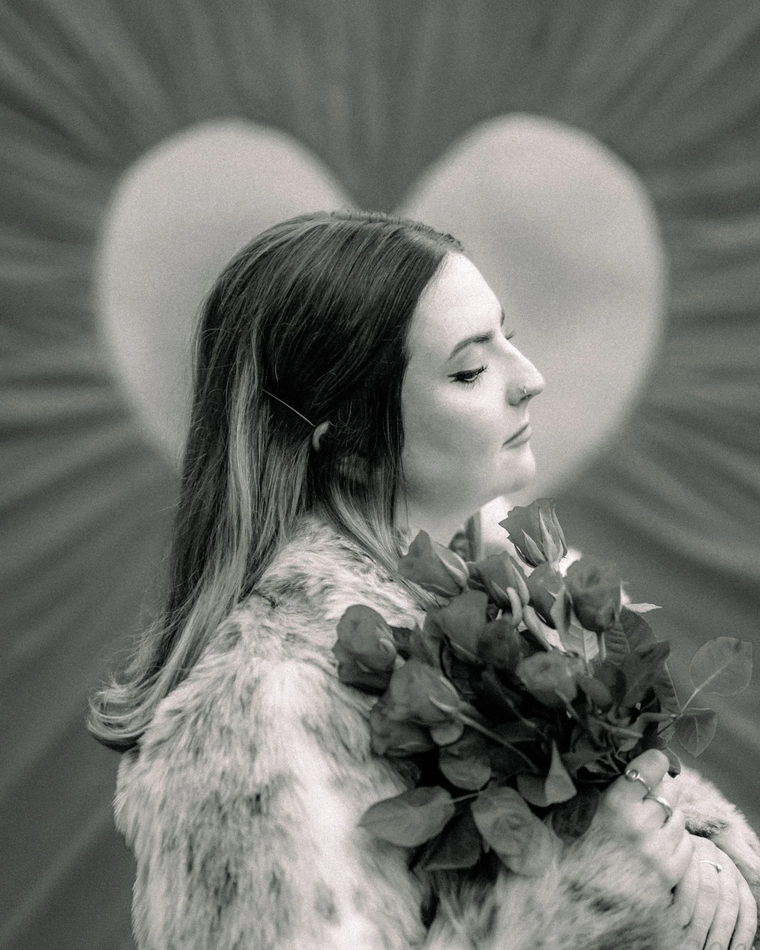 A woman with long hair, holding a bouquet of roses, is in profile with her eyes closed, in front of a heart-shaped backdrop with radiating lines.