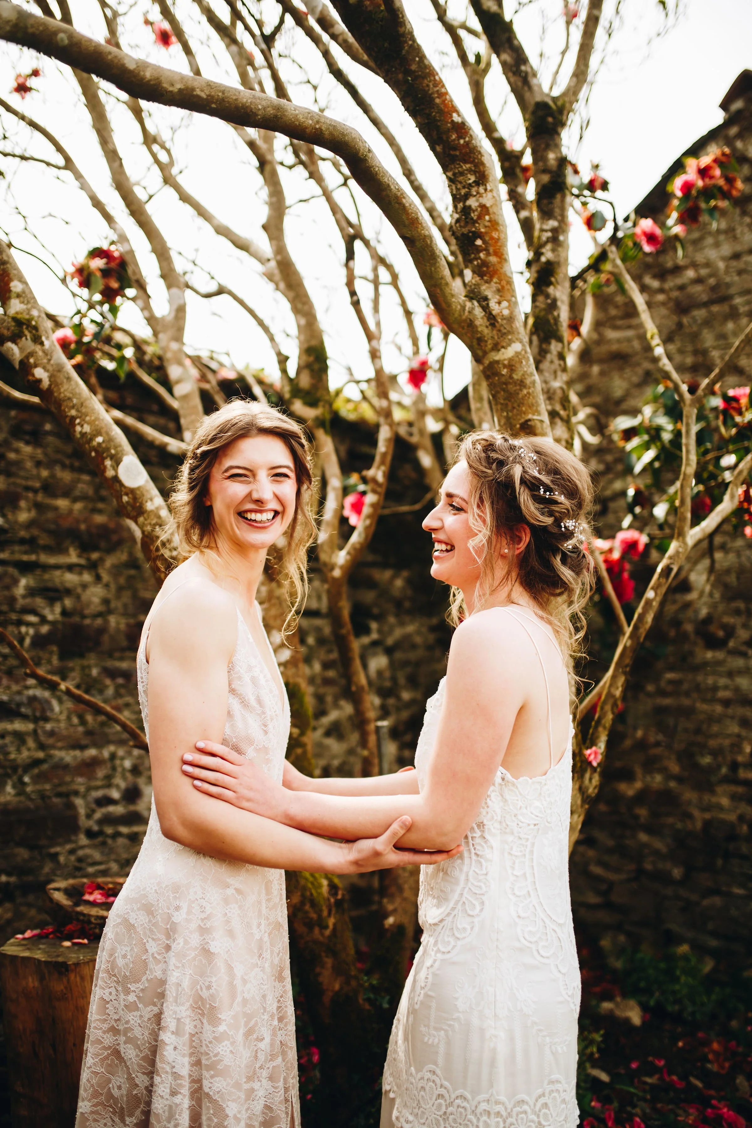 Two women in white lace dresses holding hands and laughing outdoors, surrounded by blooming tree branches and pink flowers.