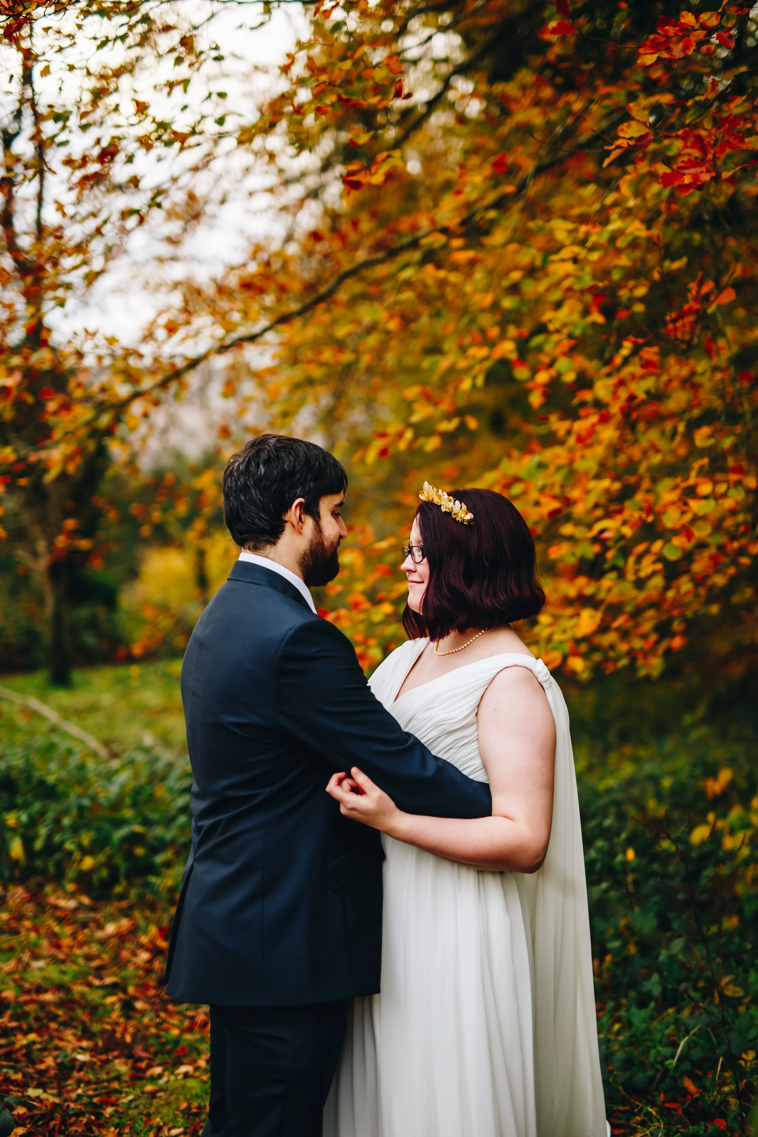 A couple dressed in wedding attire standing close together outdoors surrounded by autumnal trees with orange and yellow leaves, sharing an intimate moment.