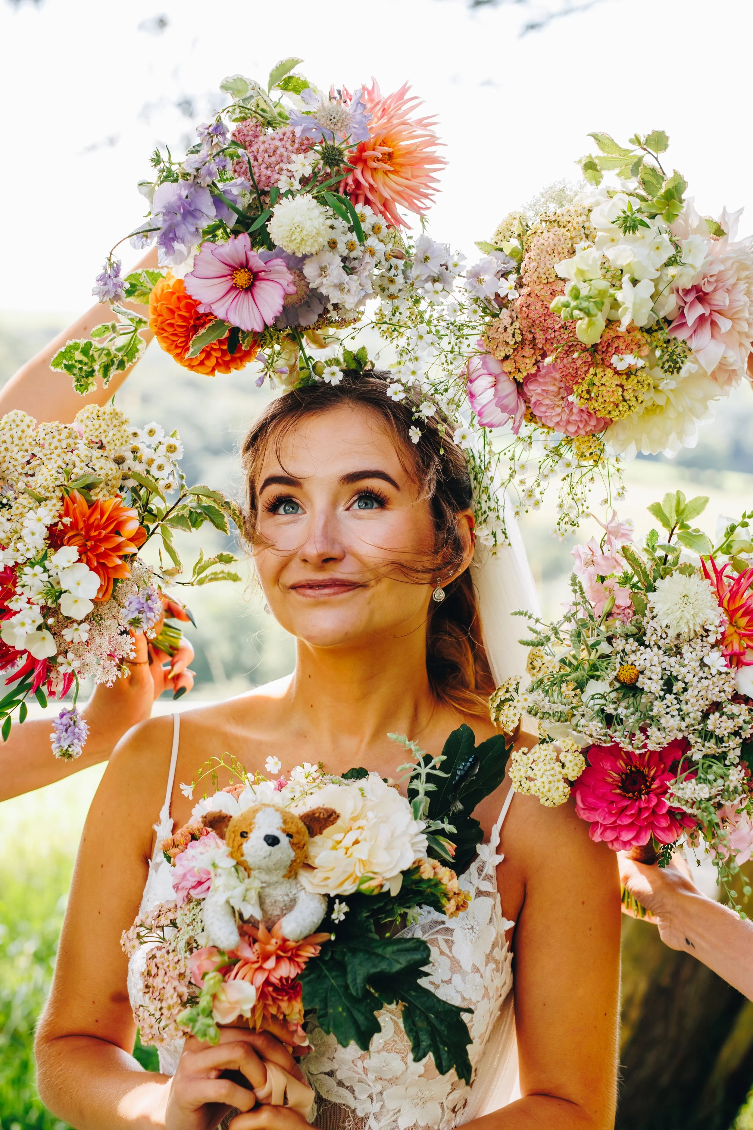Bridal portrait with a wreath of colorful flowers and a small plush toy in her hands, outdoors on a sunny day.