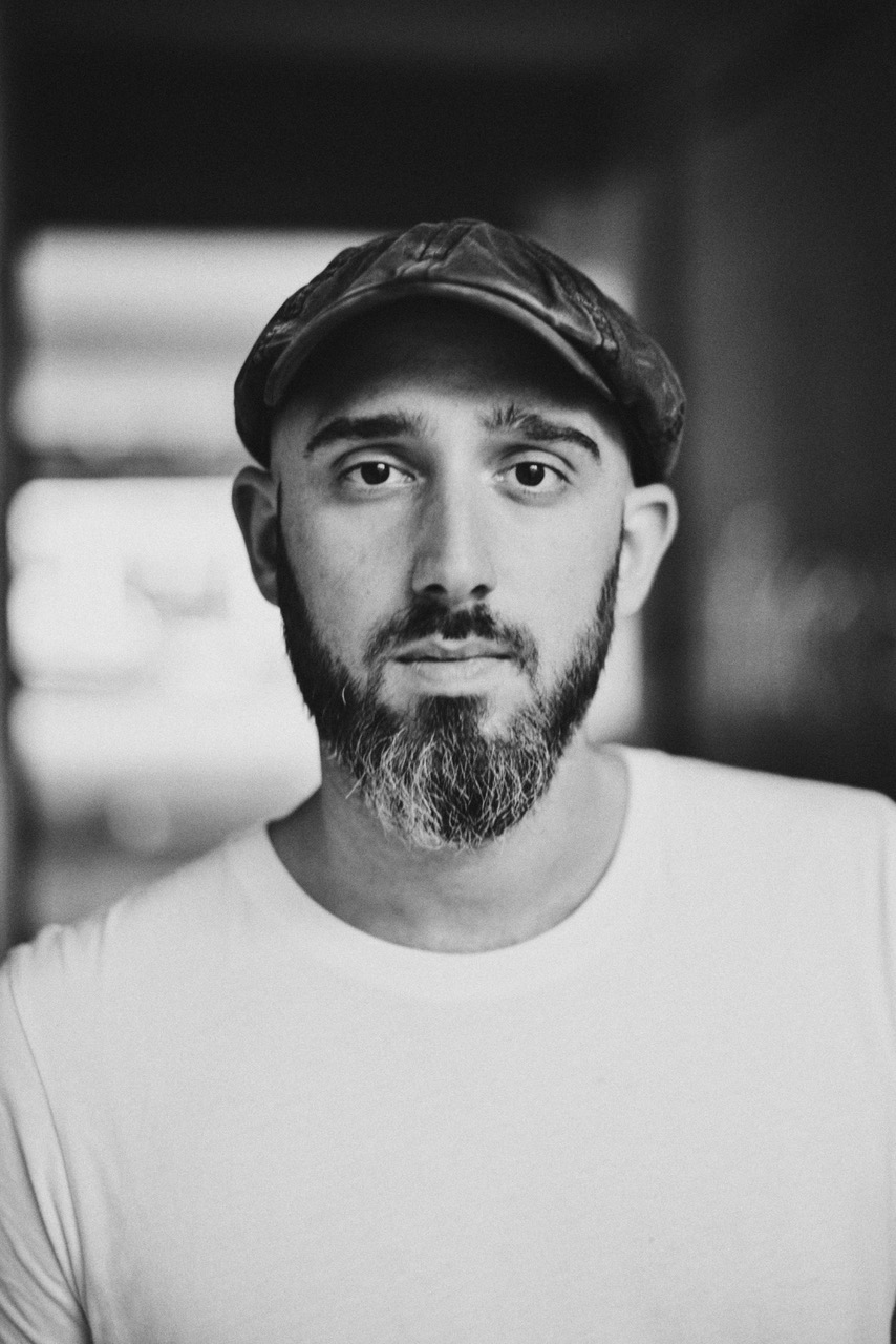 Black and white portrait of the author Yves Eugene Hensel with a beard wearing a flat cap and a white t-shirt, looking directly at the camera.